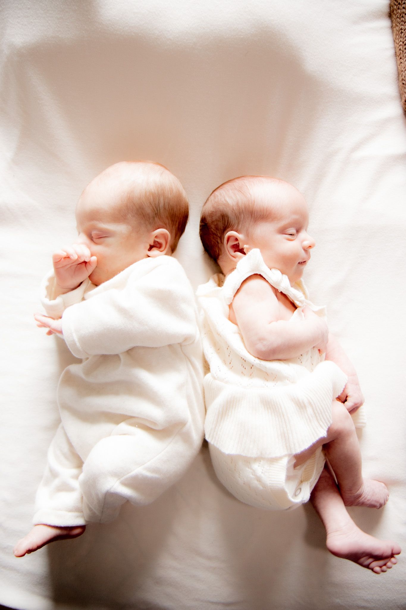 Overhead view of newborn twins sleeping back to back in white outfits on a cream blanket.