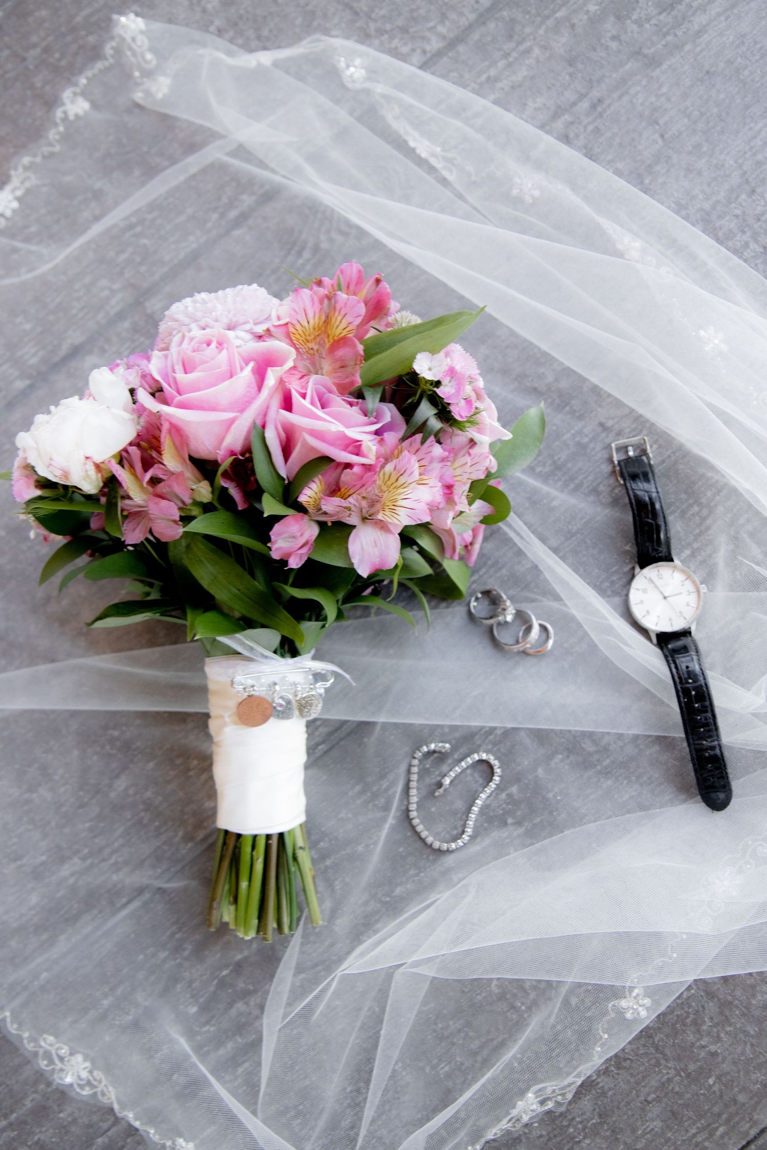 Bridal flat lay with pink rose bouquet, wristwatch, pearl bracelet, and earrings on gray surface with white lace veil.