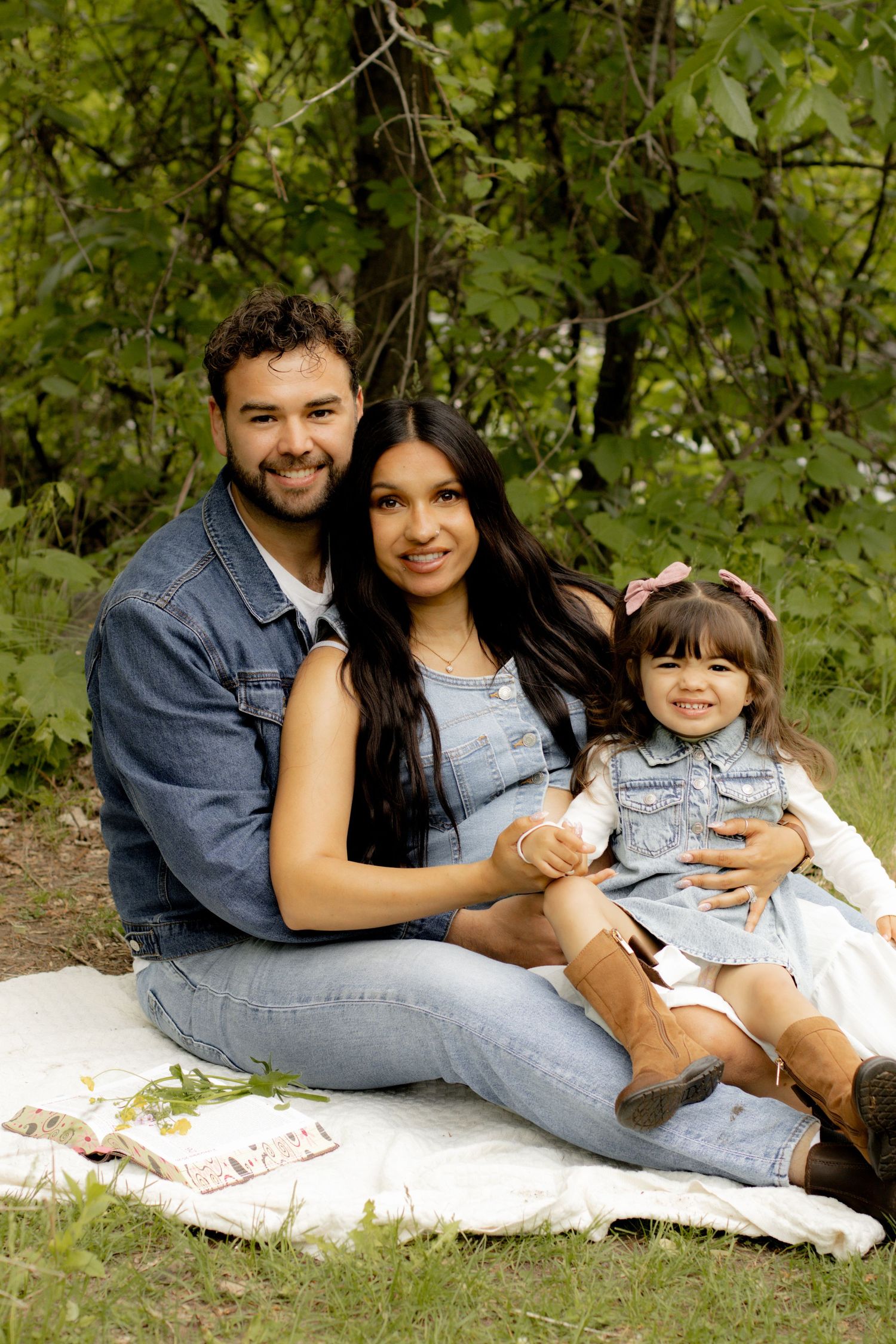 Happy family of three sitting on blanket outdoors, parents smiling with toddler girl in denim outfits among green trees.