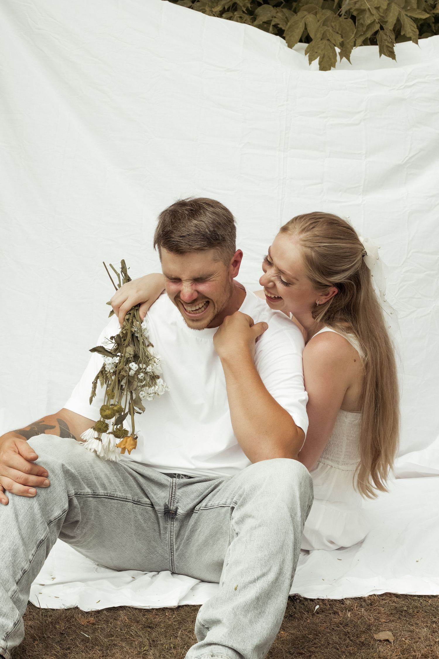 A laughing couple sitting together against a white backdrop, the woman playfully holding flowers near the mans neck.