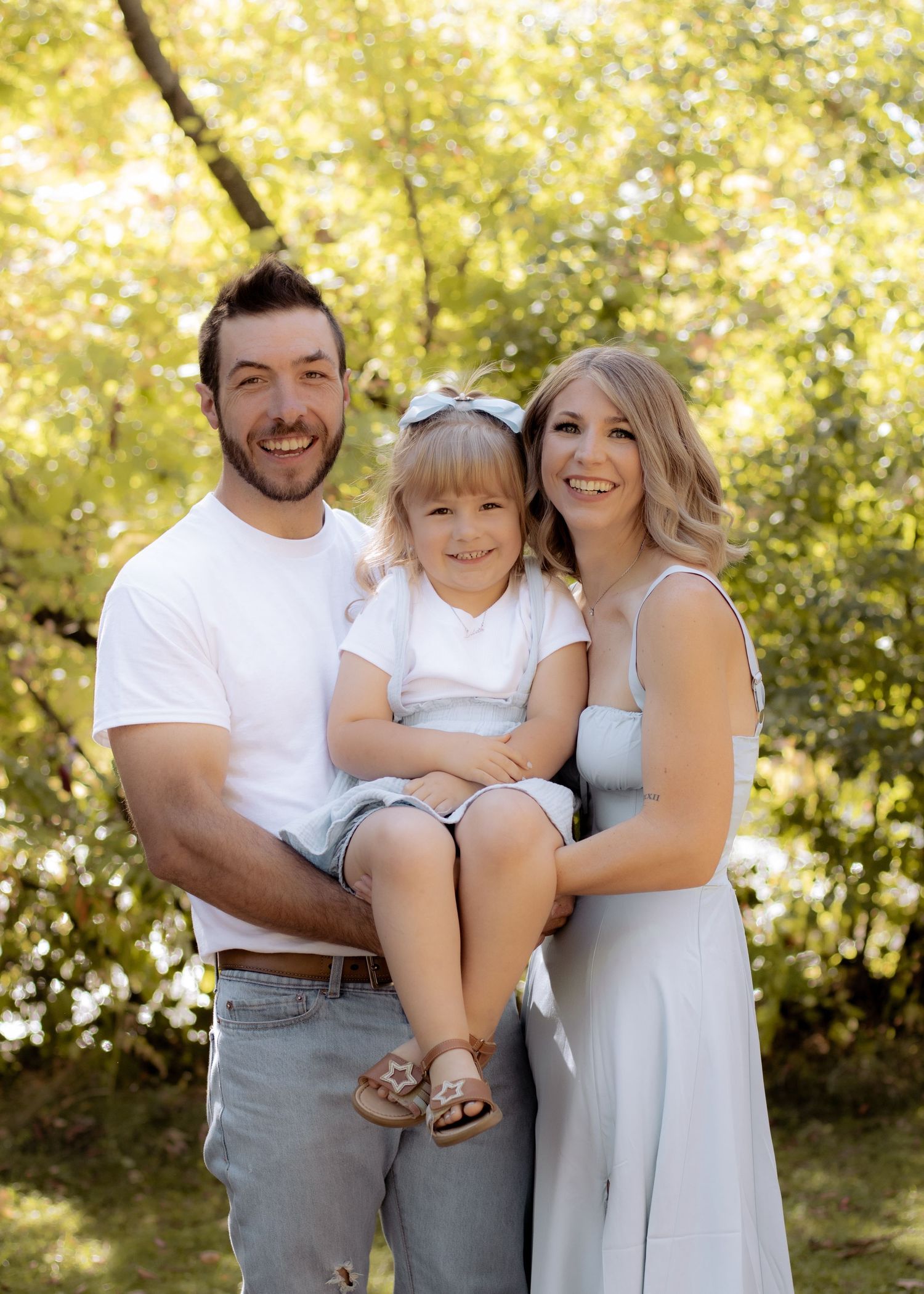 A smiling family of three poses outdoors, parents holding their young daughter among golden green trees.