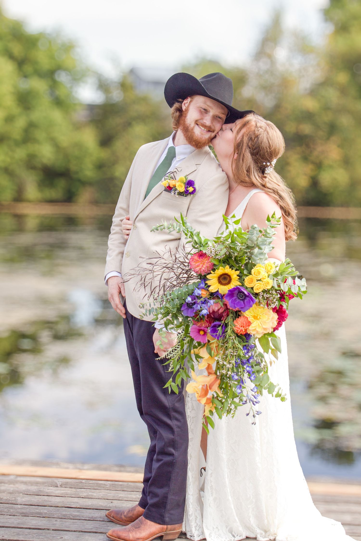 Wedding couple poses on dock with colorful wildflower bouquet and black cowboy hat.