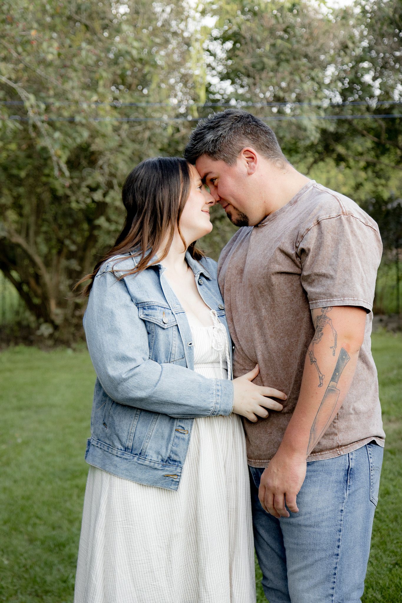 Couple sharing a romantic forehead touch outdoors, woman in white dress and denim jacket, man with tattoos in brown tee.