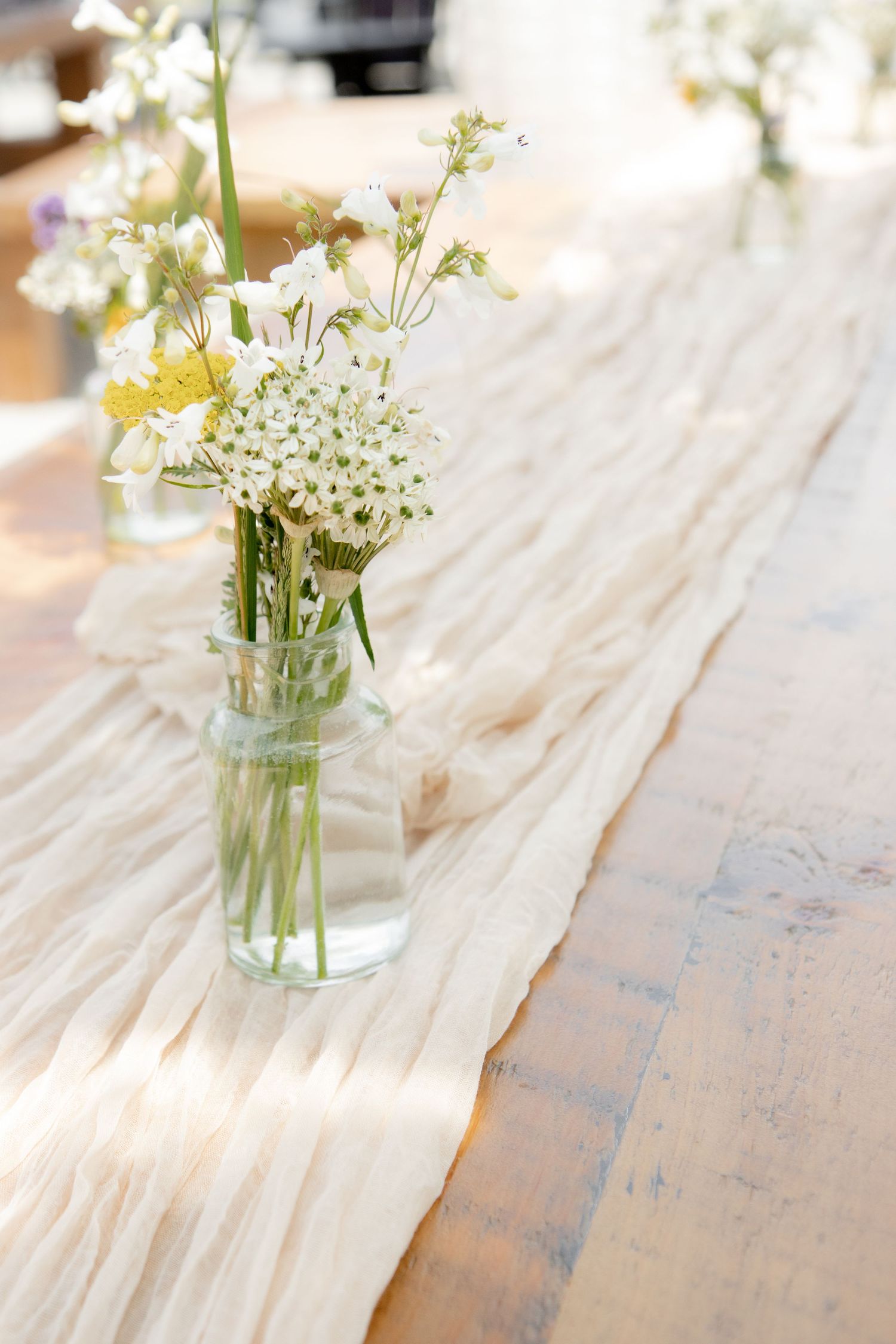 Glass jar vases with white wildflowers on a rustic wood table with a cream gauze runner in soft natural light.