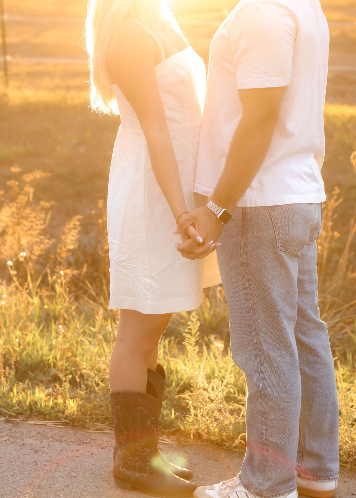 Couple holding hands at sunset on rural path, woman wearing white dress and cowboy boots.