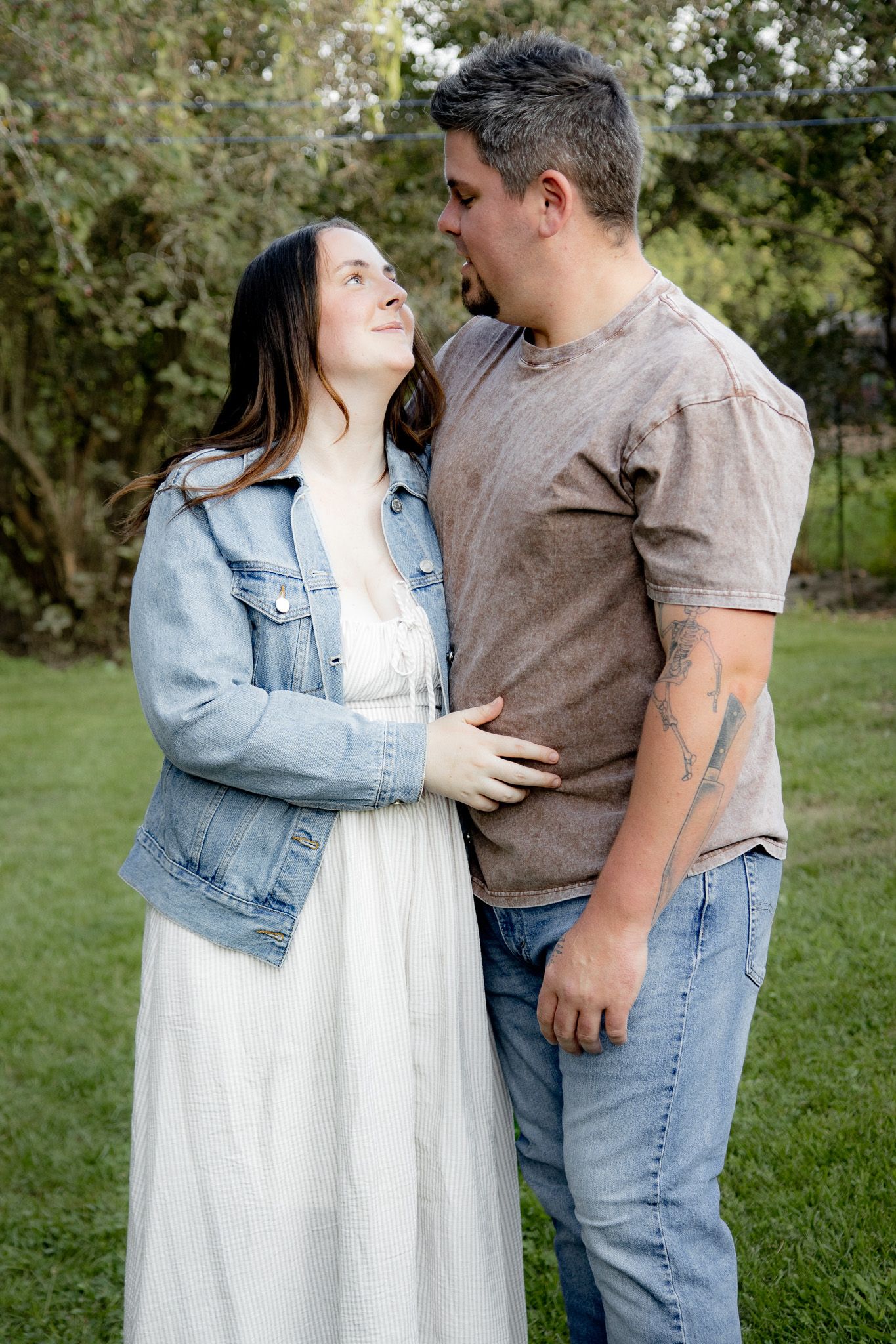A couple shares a tender moment outdoors, woman in white dress and denim jacket, man in brown tee and jeans.