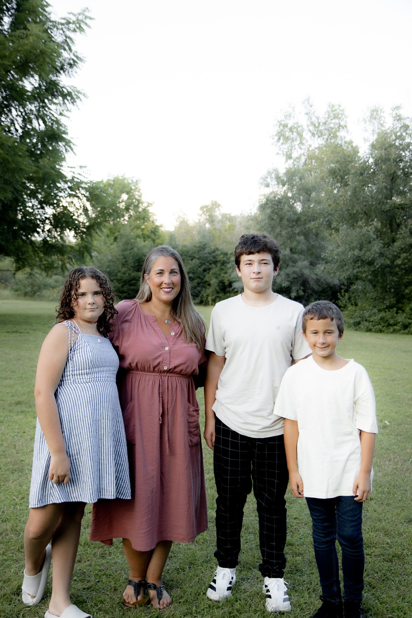 A smiling family of four poses outdoors on green grass with trees in the background on a bright day.