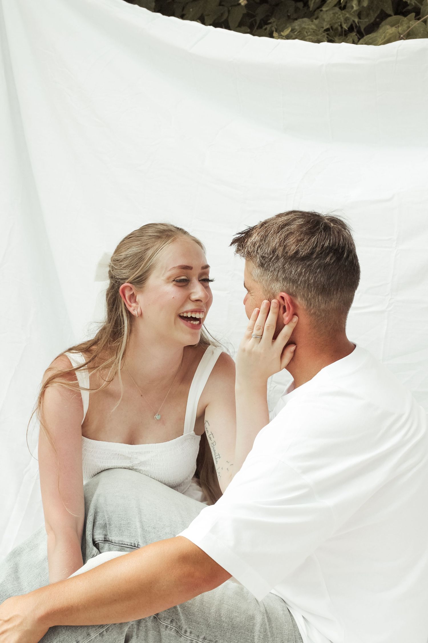 Laughing blonde woman touches mans face while sitting together against white fabric backdrop.