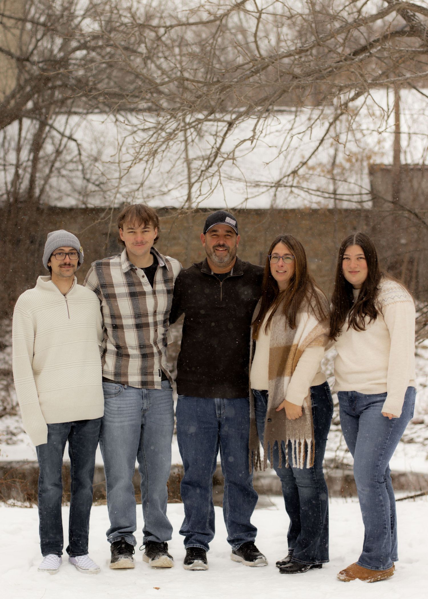 Family of five posing together outdoors in snow, wearing casual winter clothing, smiling at camera.