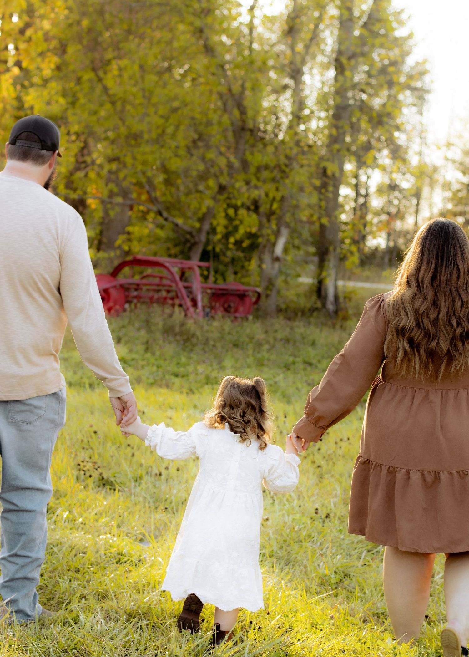 Parents hold hands with young girl in white dress walking toward red farm equipment in autumn meadow.