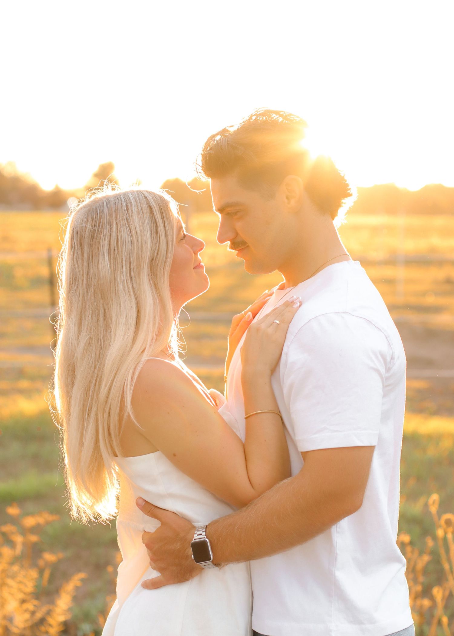 Romantic couple embracing at golden sunset in open field, woman in white dress.