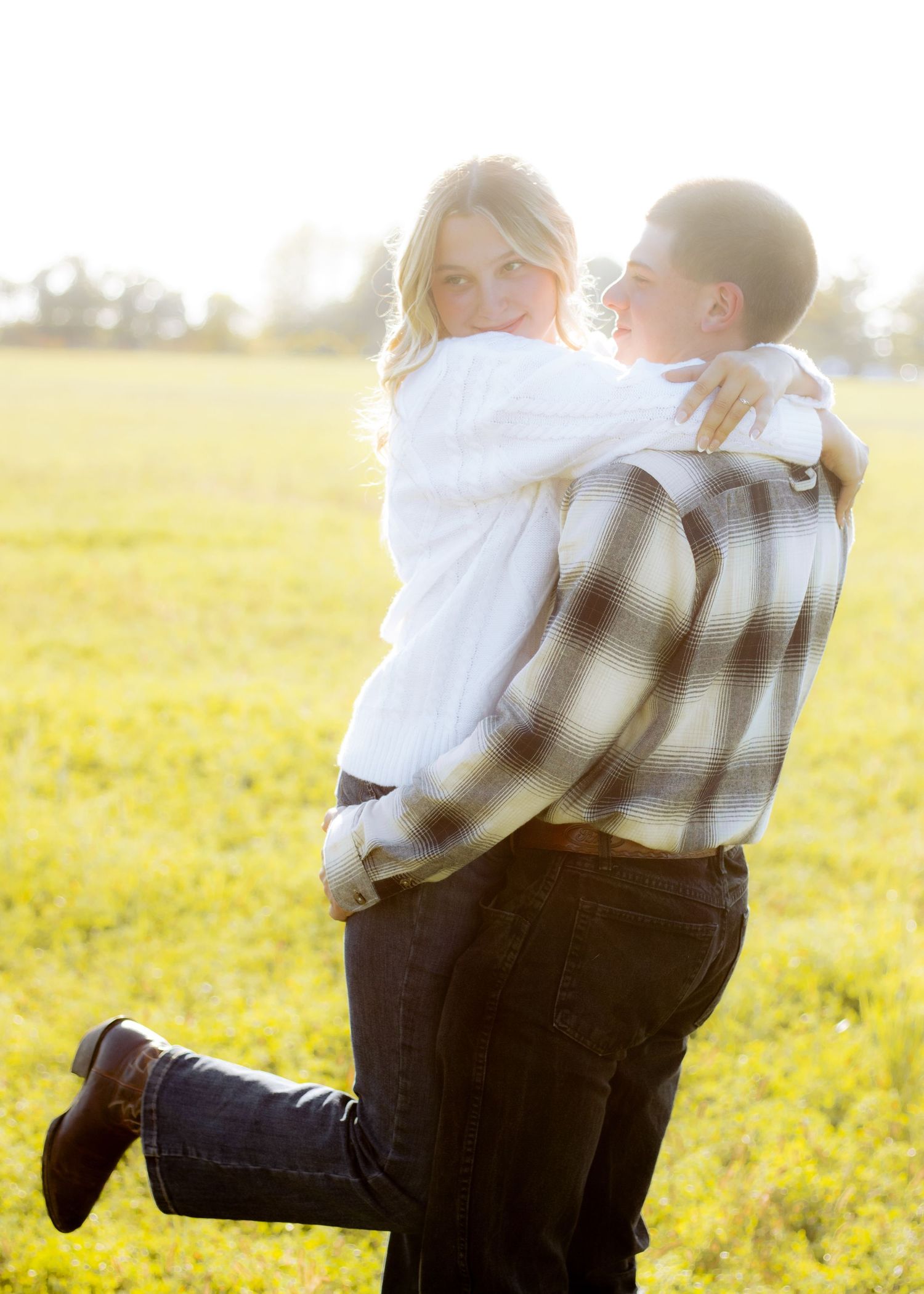 Couple embracing in a sunlit green field, woman in white sweater smiling at camera while man in plaid shirt lifts her.