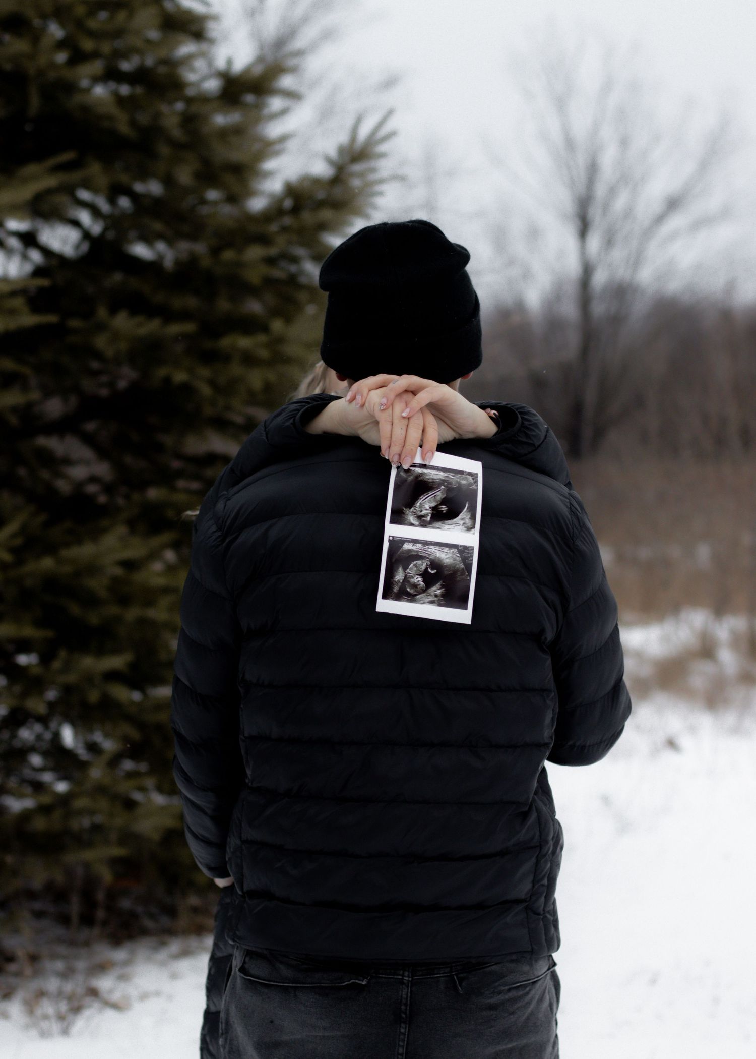 Person in black jacket holding ultrasound photos behind their back in snowy winter setting.