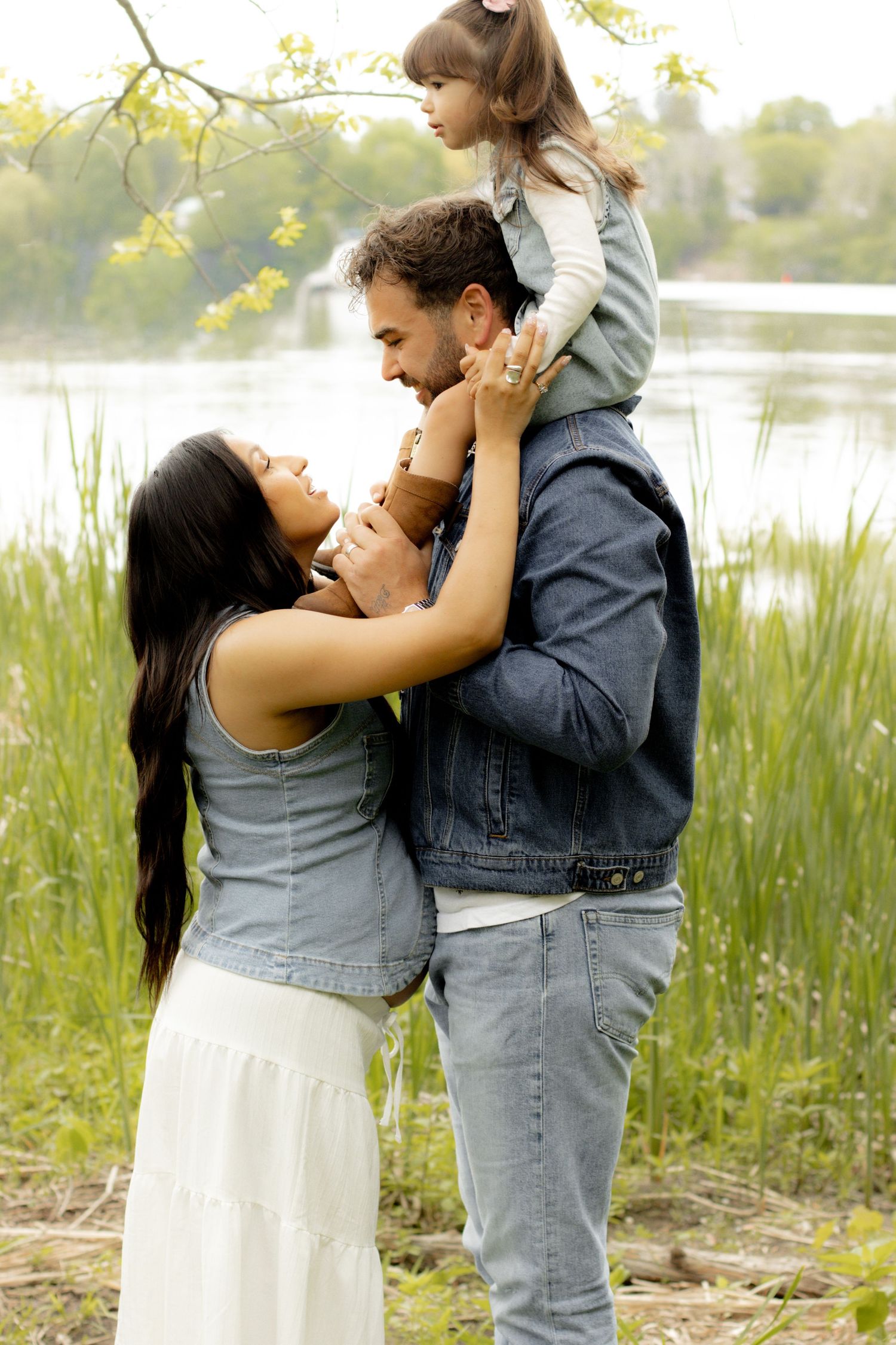 A happy family of three enjoying time outdoors near a lake, with dad carrying a toddler on his shoulders.