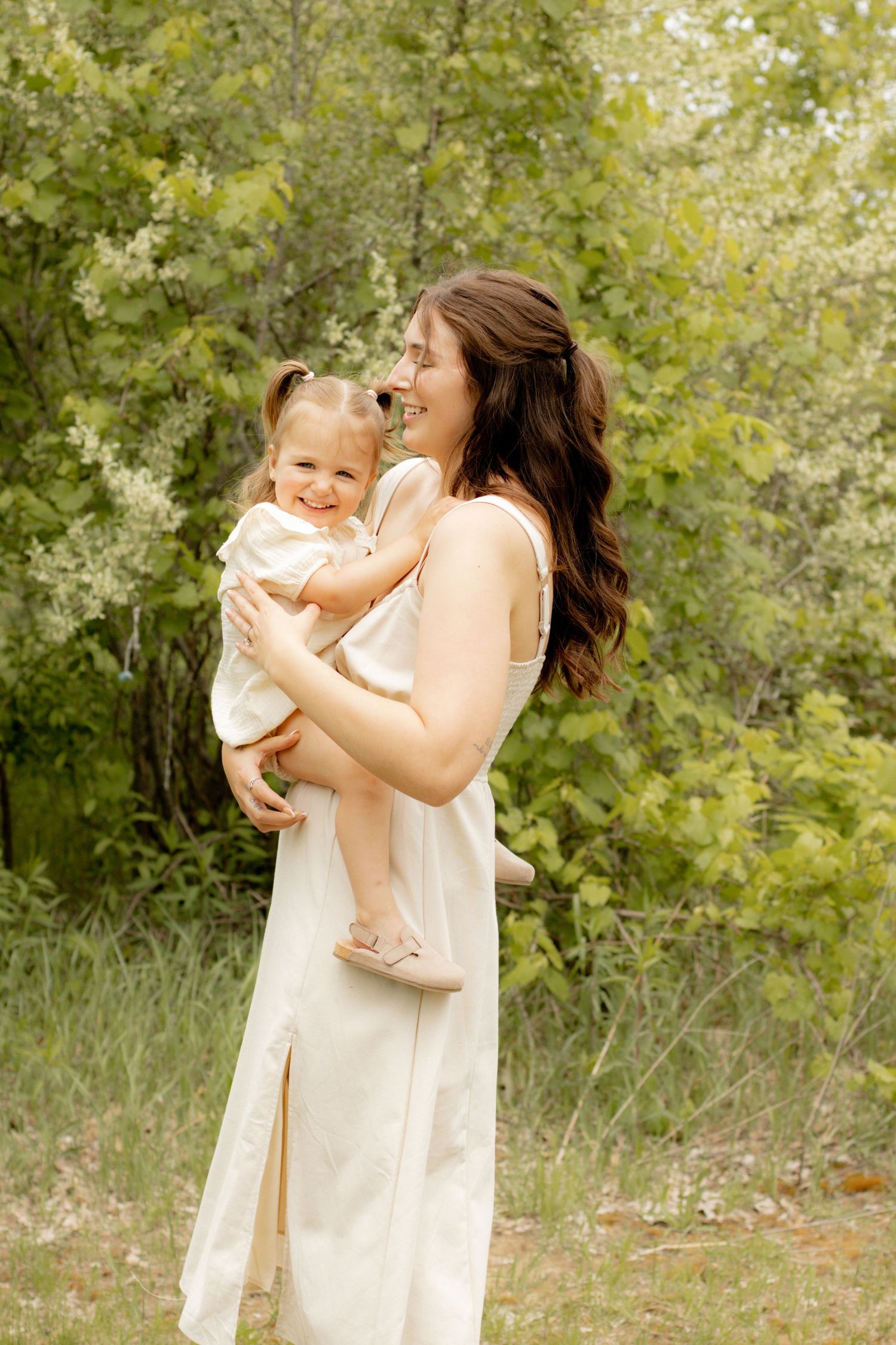 A laughing mother in a white dress holds her smiling toddler daughter outdoors among lush green trees.