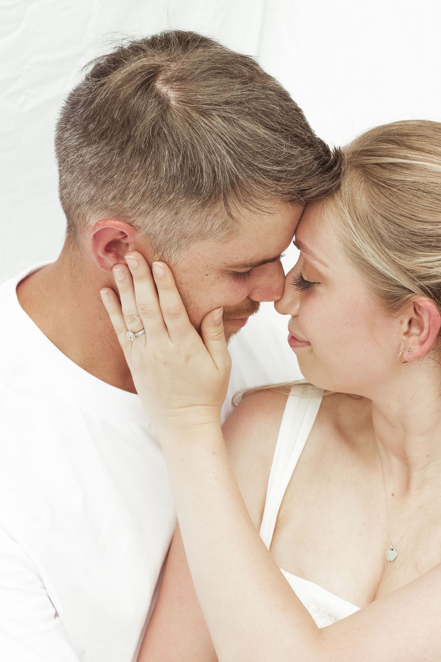 Intimate close-up of couple touching foreheads, woman wearing engagement ring in soft white lighting.