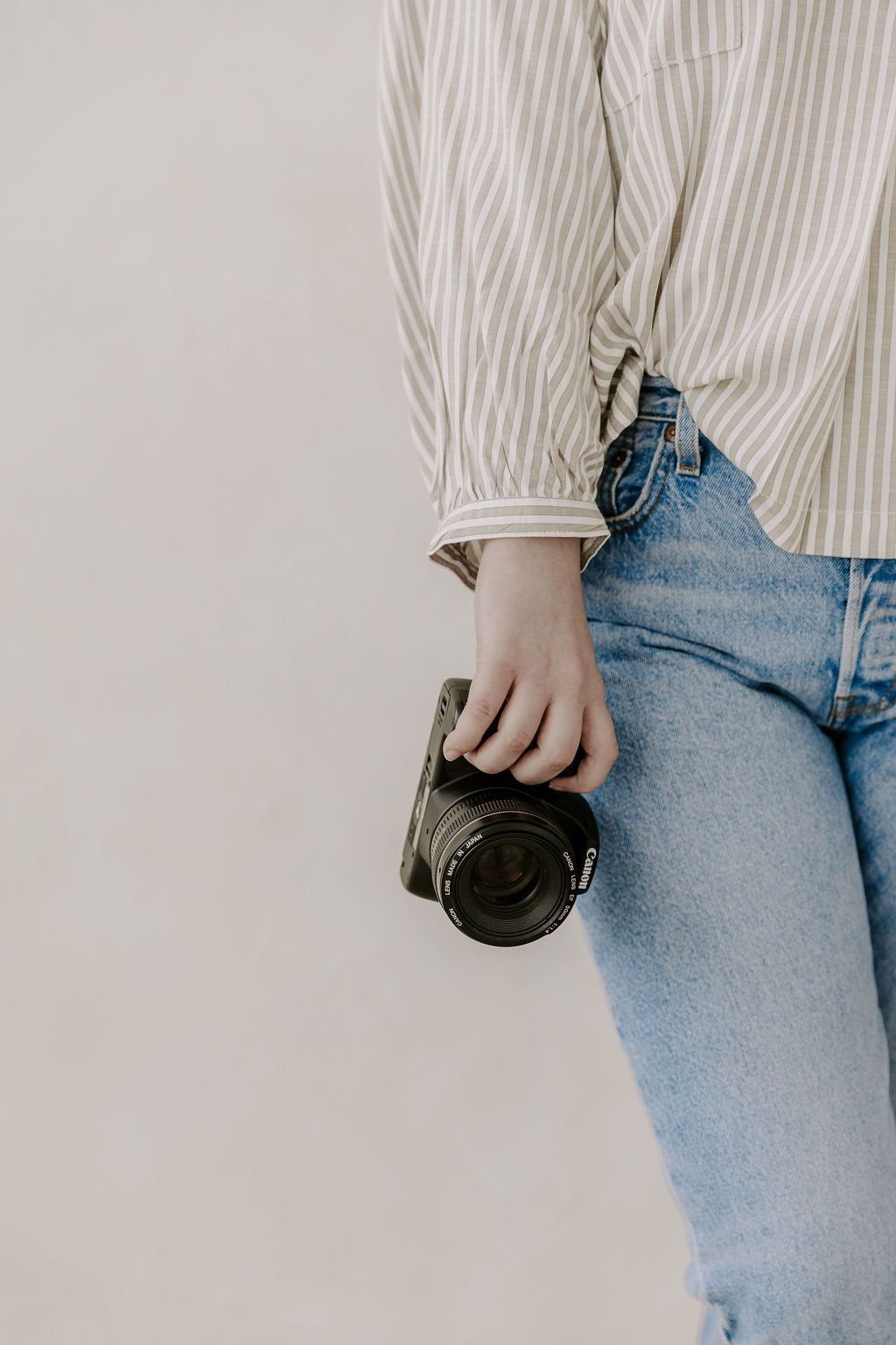 Close-up of a person in striped shirt and jeans holding a black camera against a neutral wall.