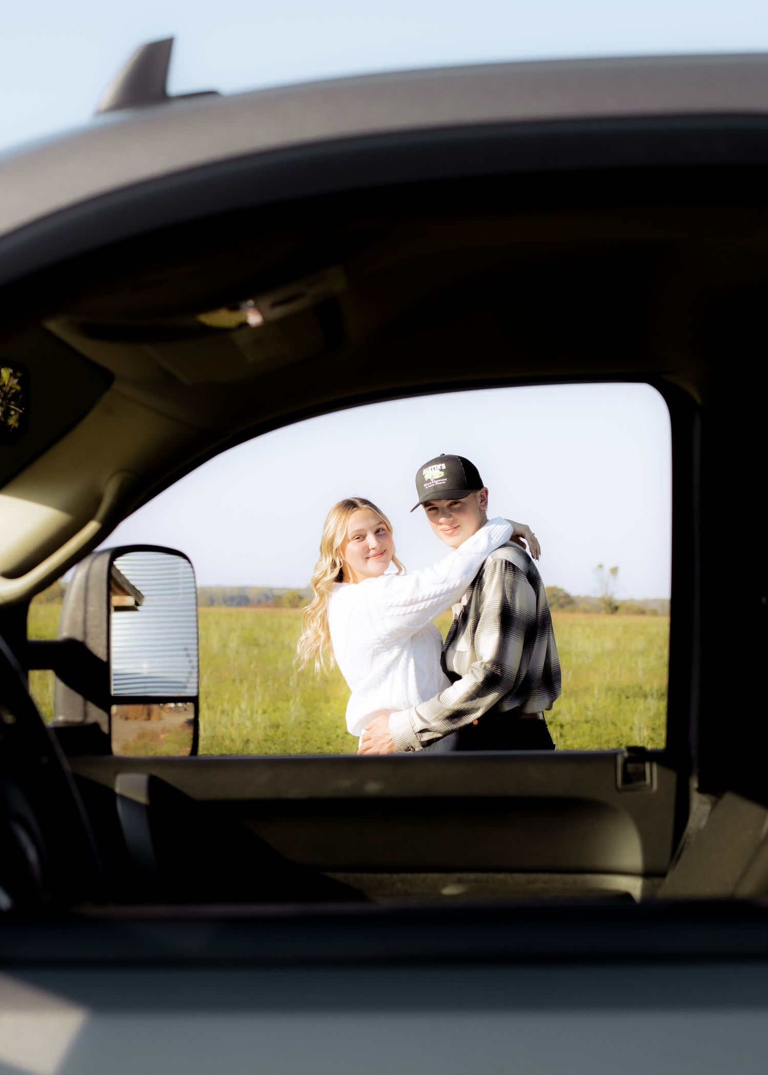 A couple embraces in a green field, photographed through the window of a truck in golden hour light.