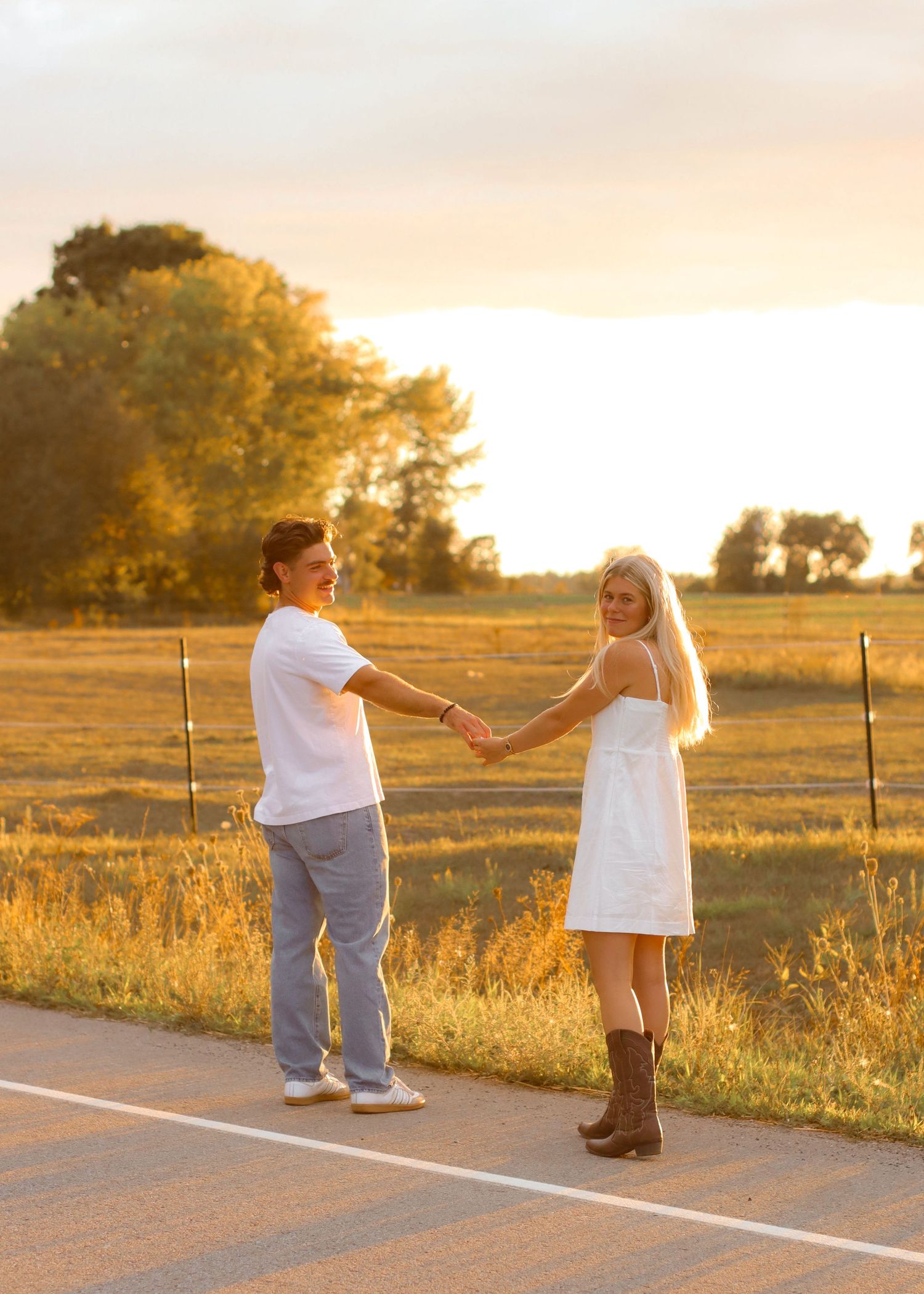 Couple holding hands on rural road at sunset, woman in white dress and cowboy boots.
