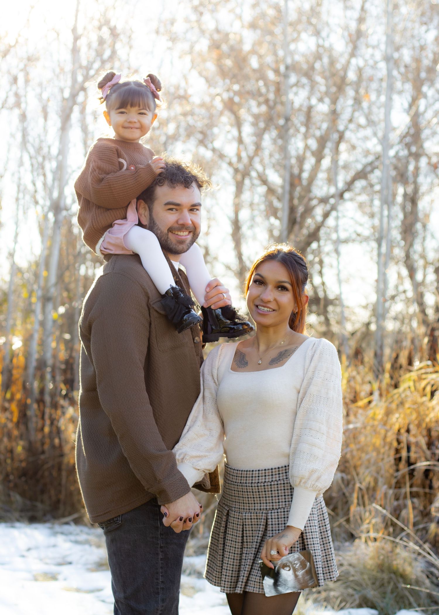 Family portrait with parents and toddler daughter on dad's shoulders in a snowy outdoor winter setting.