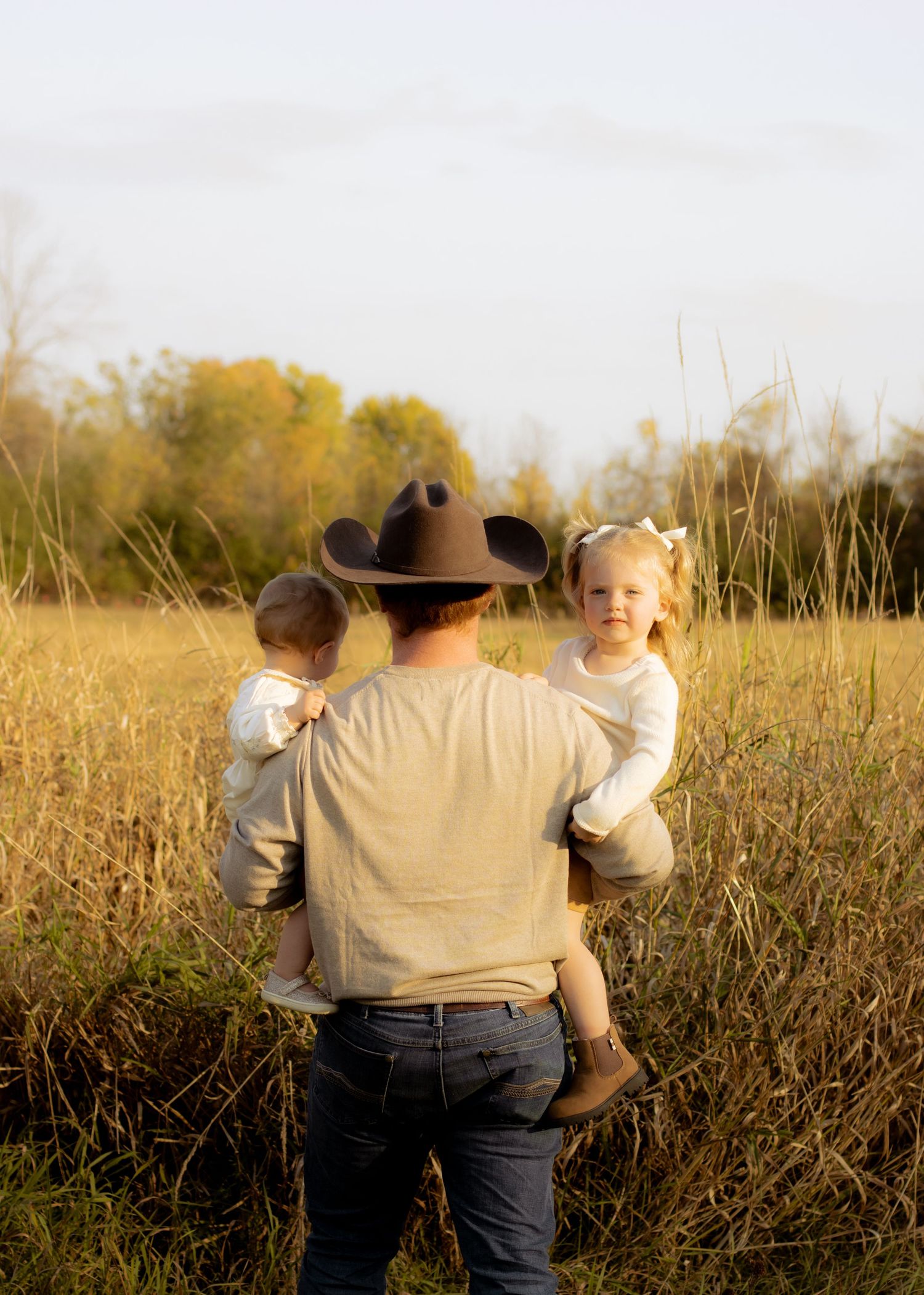 Father in cowboy hat carries two young children through a golden field at sunset.