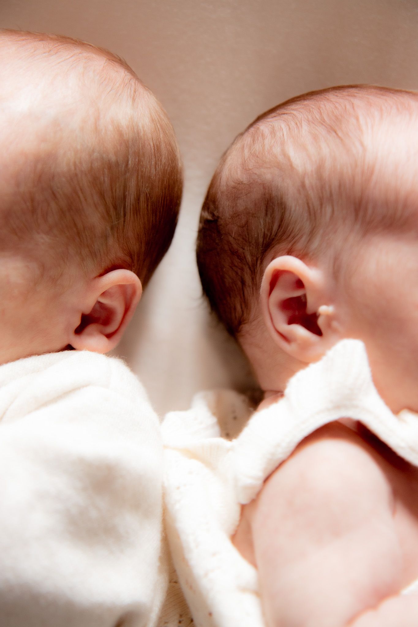 Two newborn twins lying side by side, seen from above, wearing white onesies with fine hair visible.