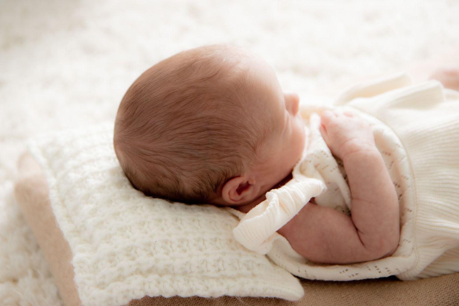 Newborn baby sleeping peacefully on a white knit blanket, wearing a cozy white outfit.