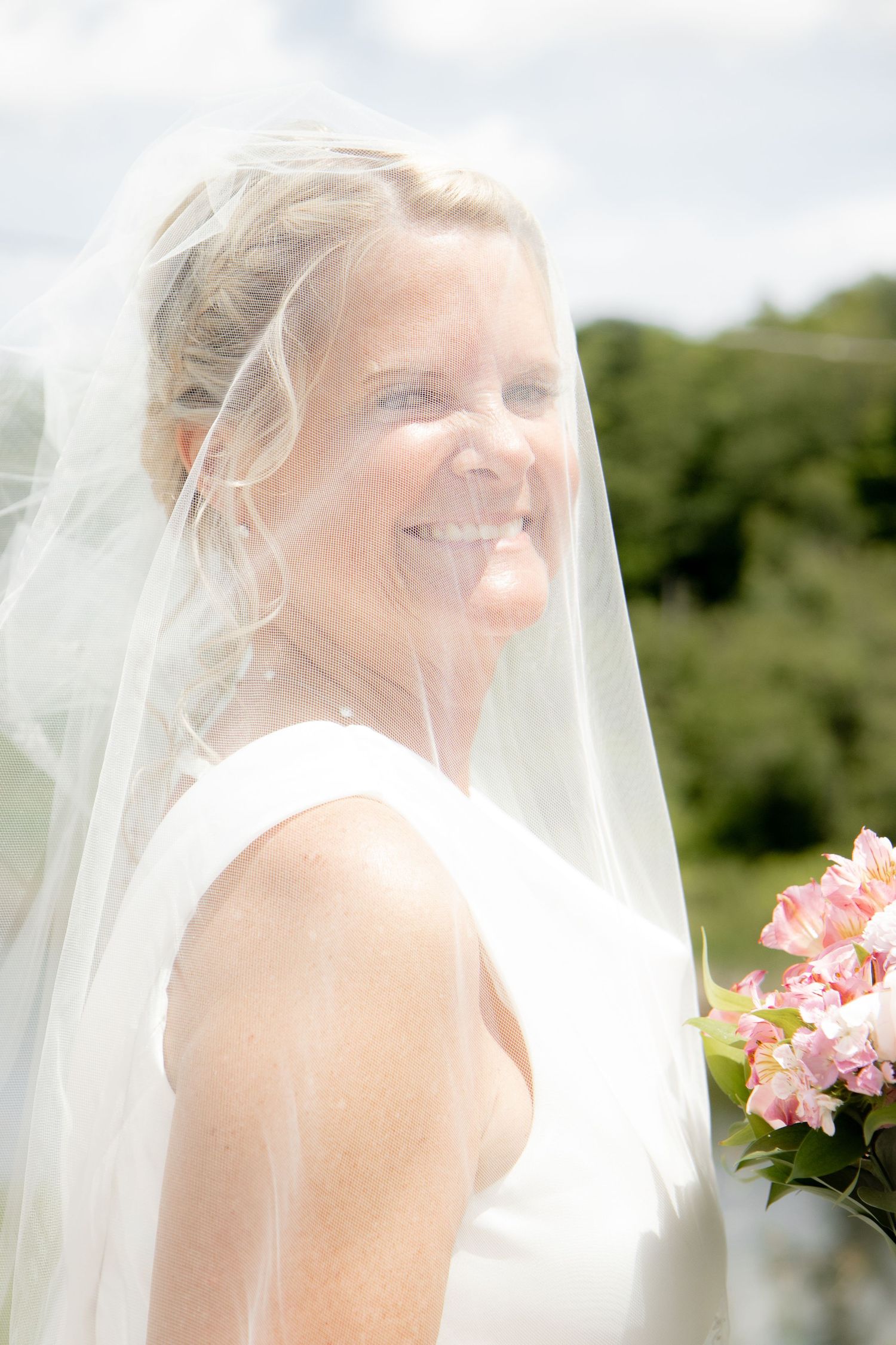 Smiling bride in white dress and veil holding pink bouquet outdoors on sunny day.