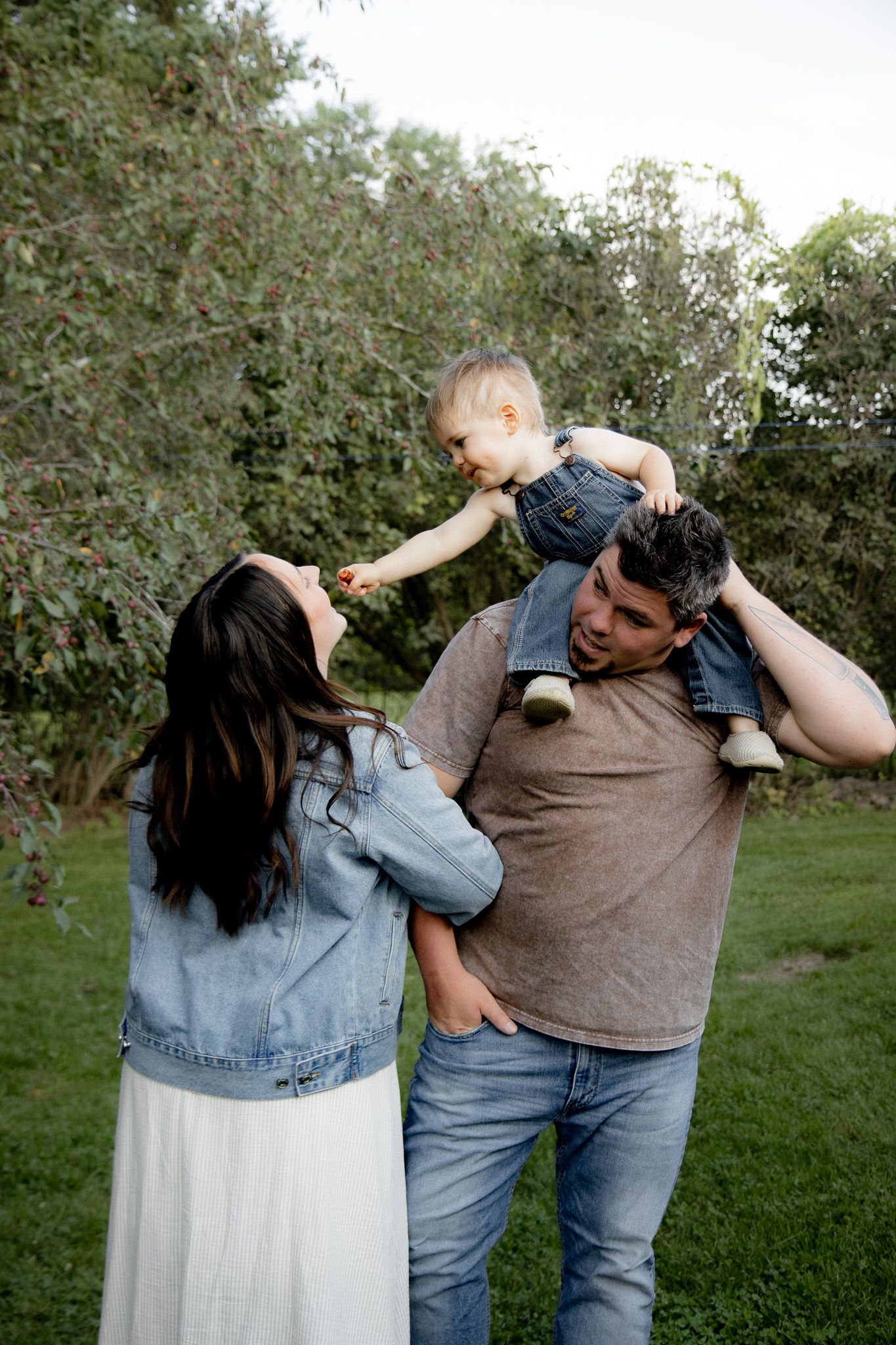 A happy family with a baby perched on dad's shoulders outdoors in a lush green garden setting.