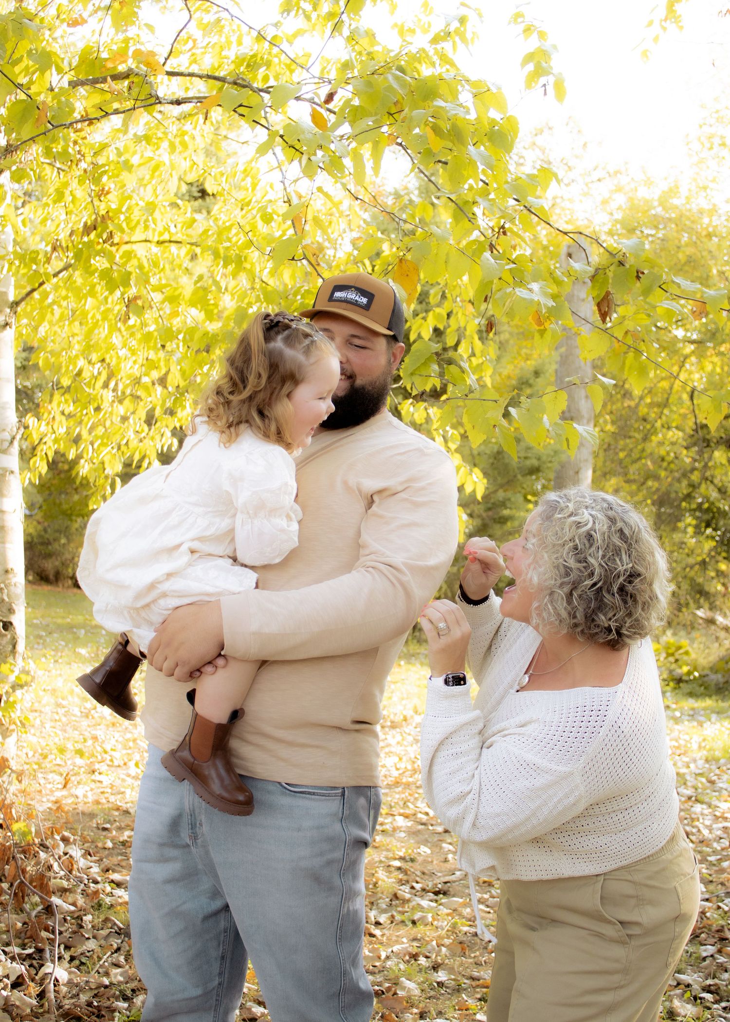 Bearded father holds toddler girl while grandmother playfully interacts with child outdoors in autumn setting.