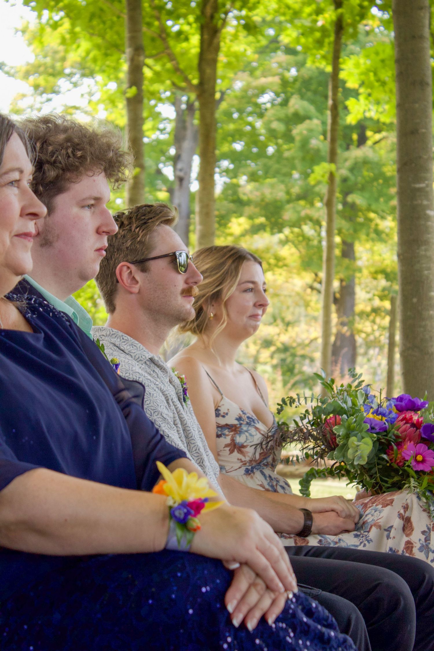 Wedding guests seated outdoors in wooded ceremony holding colorful floral bouquet.