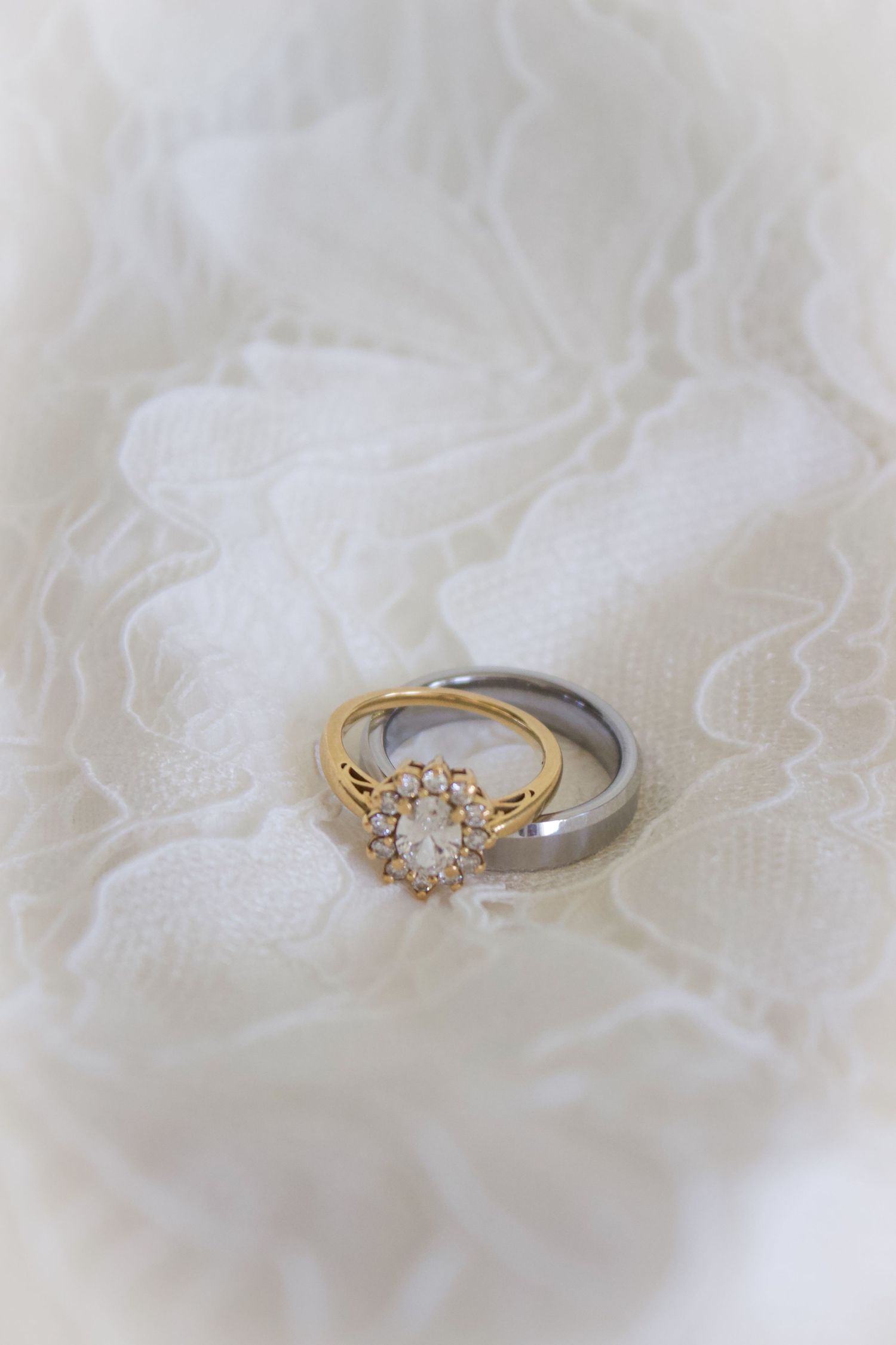A gold diamond halo engagement ring paired with a silver wedding band resting on white lace fabric.