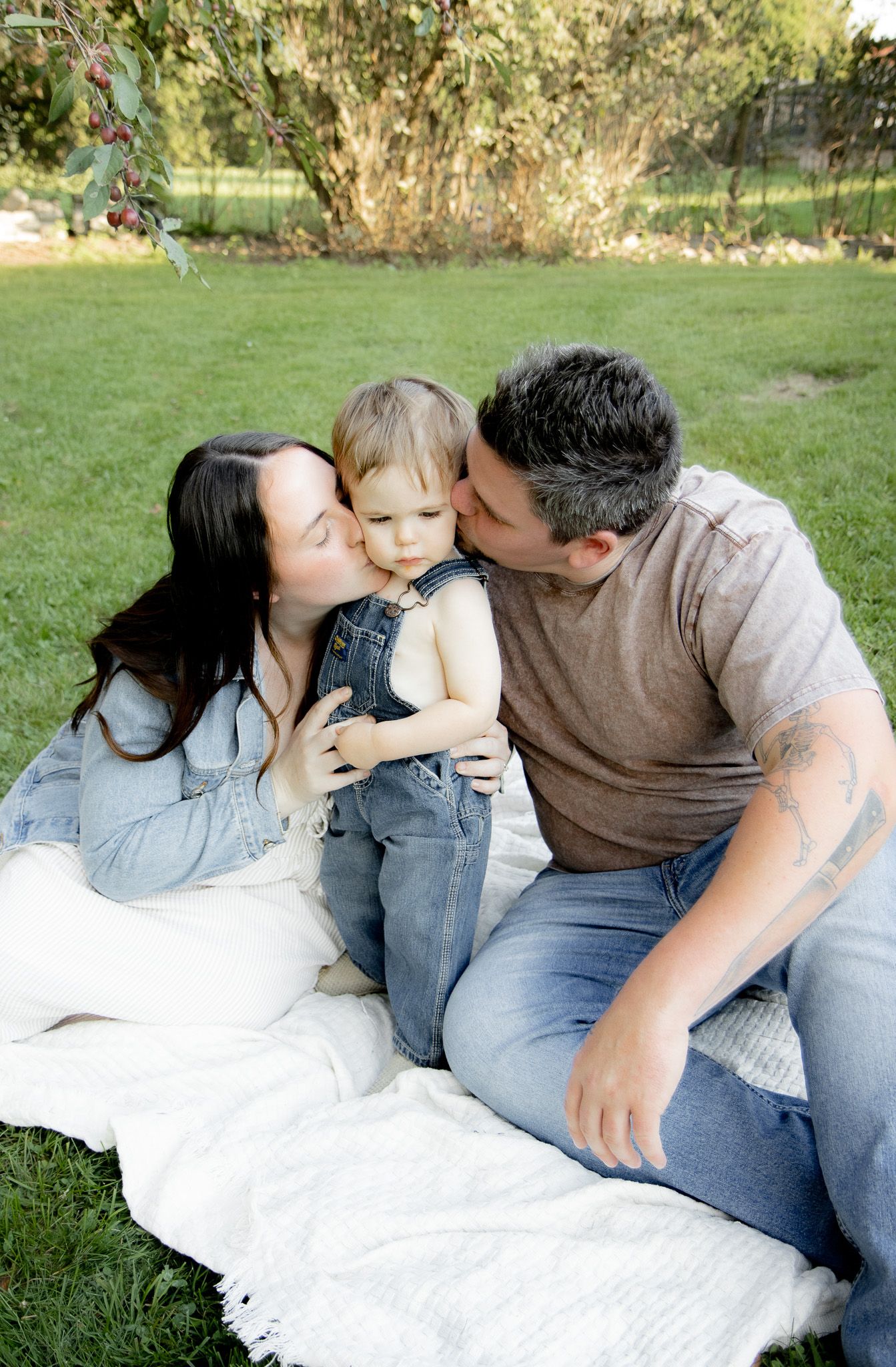 Parents kissing their toddler on cheeks while sitting on white blanket in green backyard.
