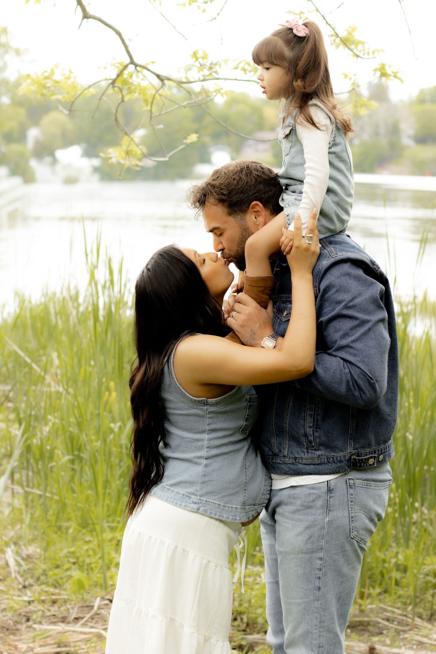 A happy family with parents kissing while father holds young daughter on shoulders near a scenic lake.