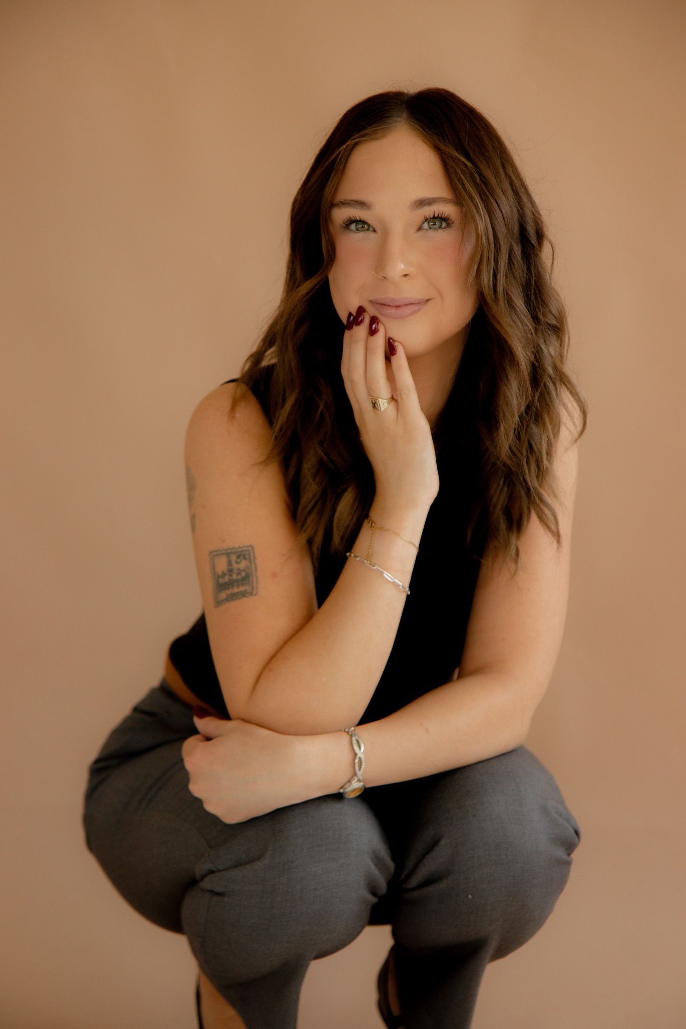 Young woman with wavy brown hair poses confidently in black sleeveless top and gray pants against beige background.