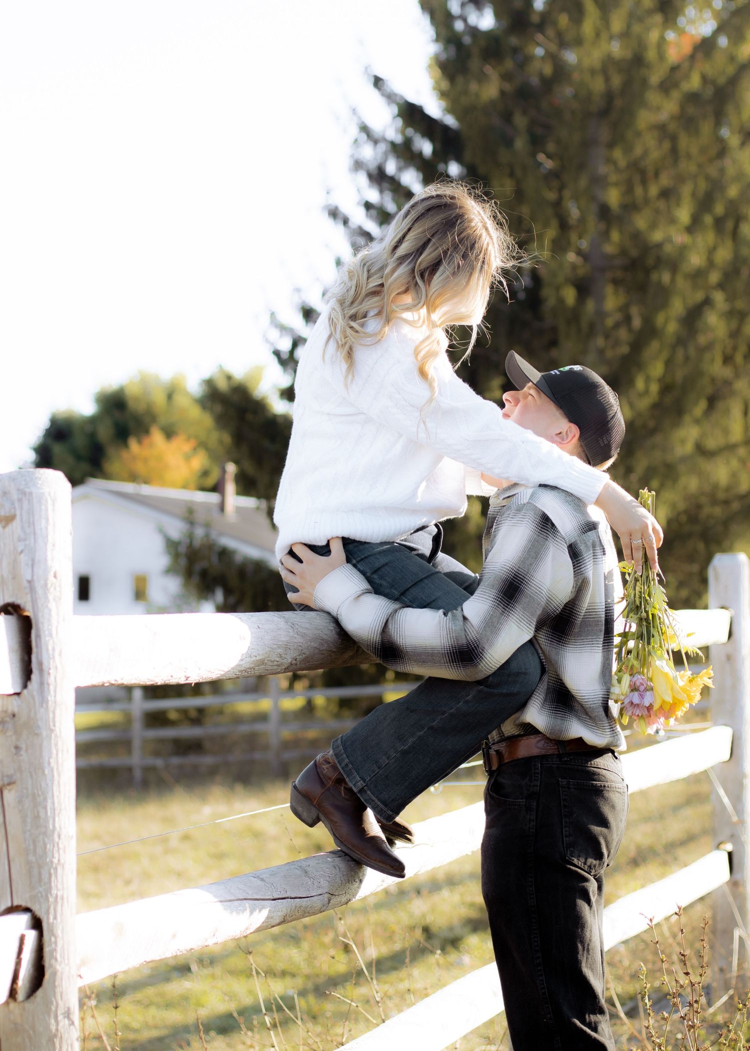 Man in plaid shirt lifting woman in white sweater sitting on a white wooden fence at a sunny farm.
