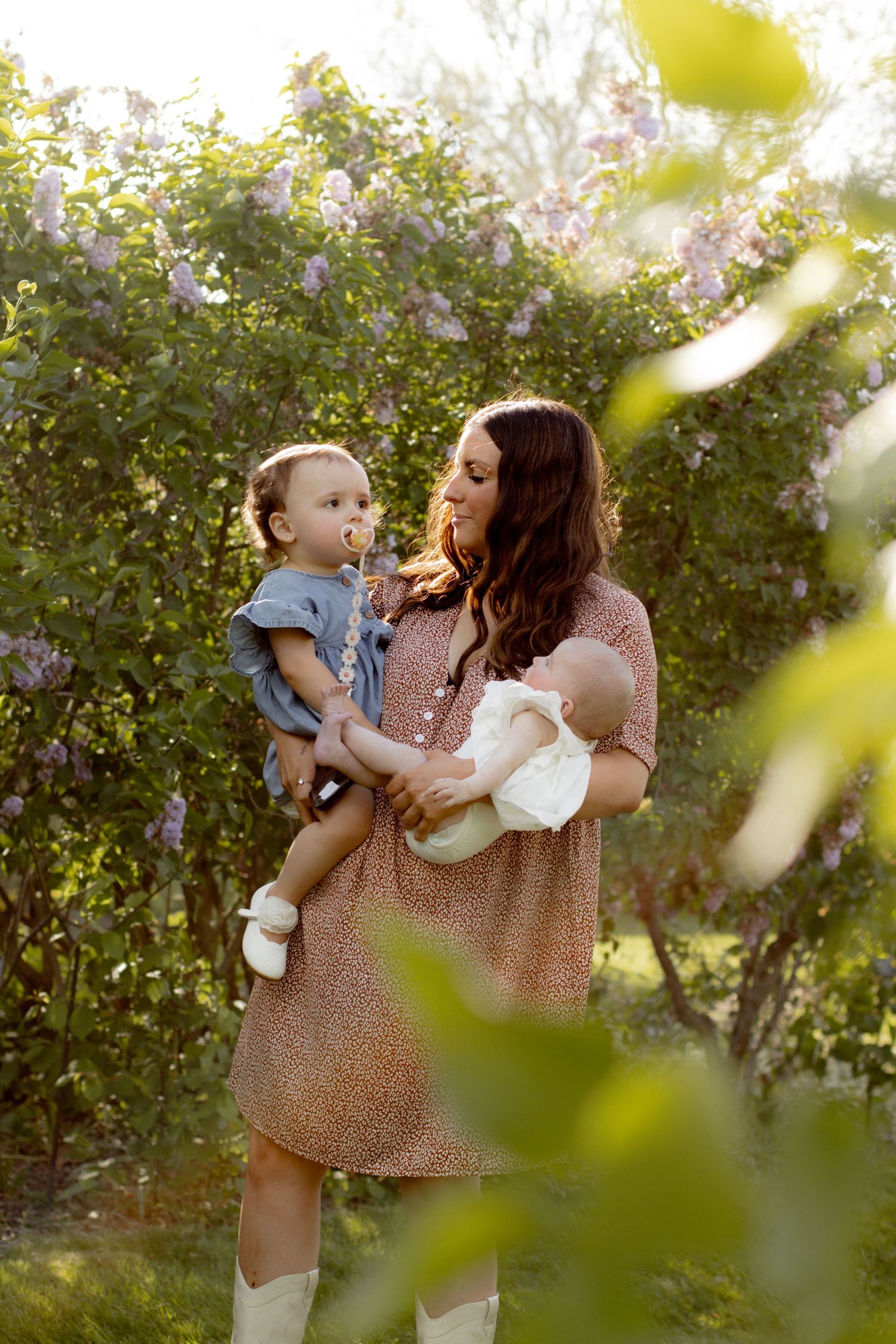 Young mother in floral dress holds toddler and newborn baby among blooming lilac bushes.