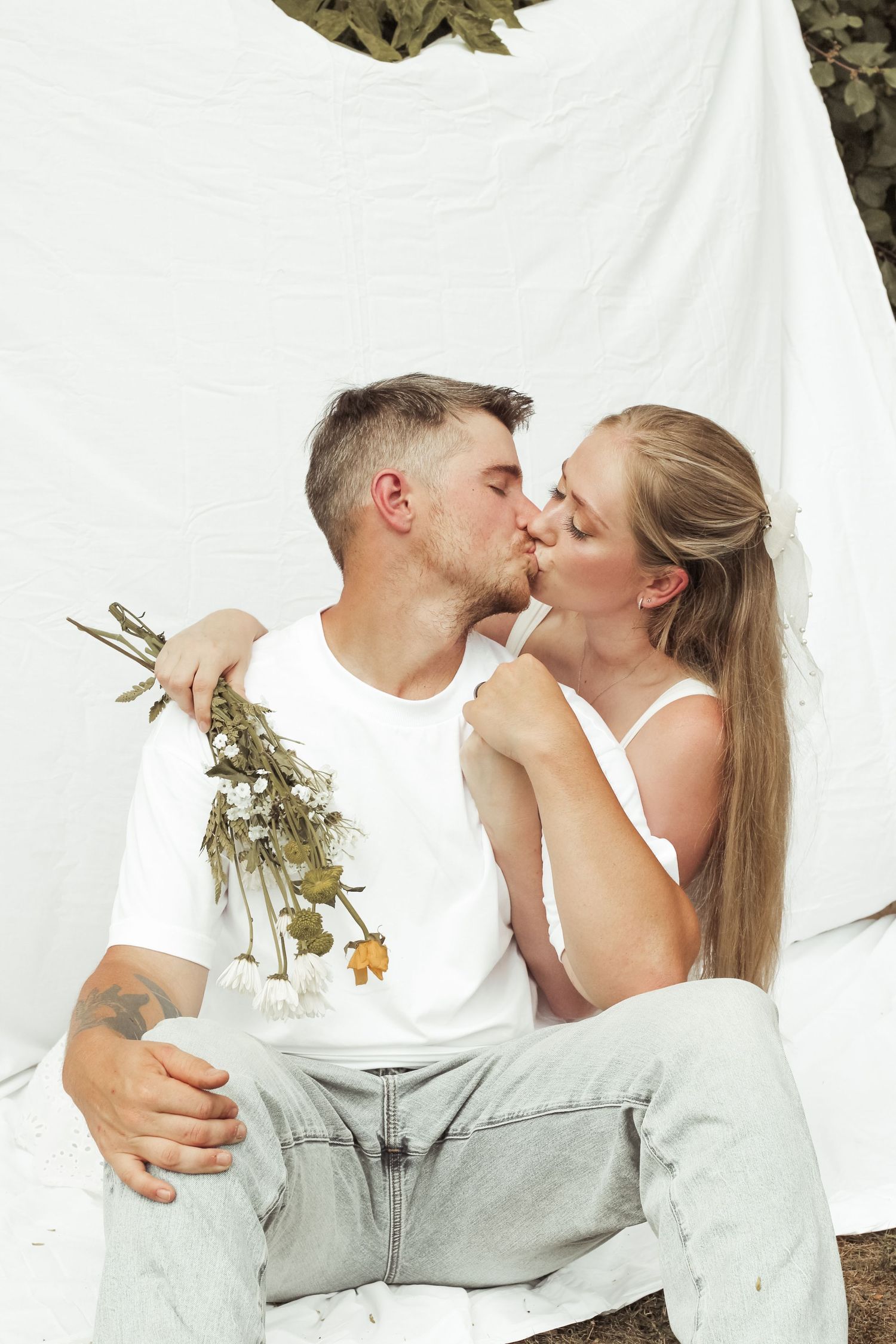 Couple kissing while seated outdoors against a white fabric backdrop, man holding dried wildflowers.