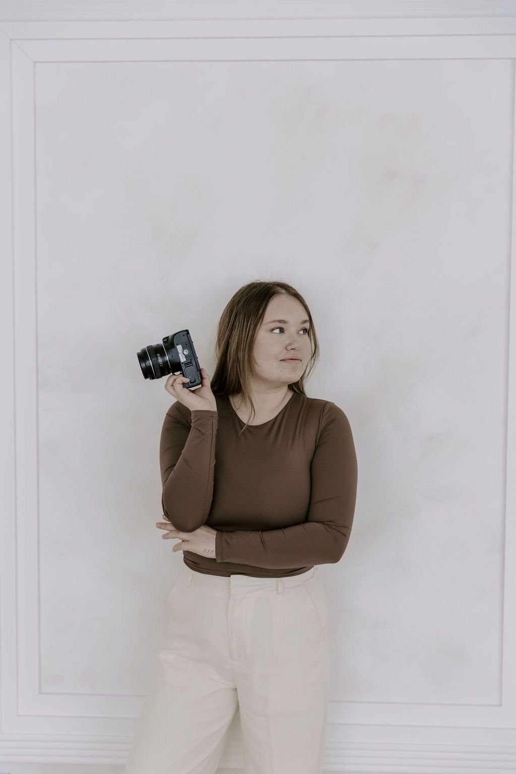 Young female photographer holding camera against white wall in brown long-sleeve top.
