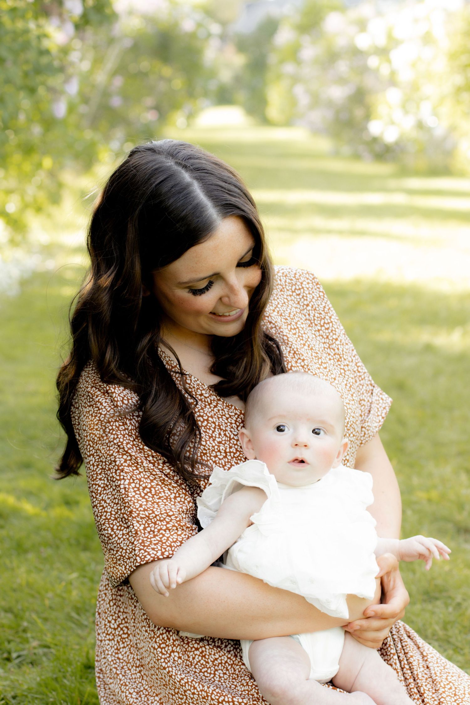 A dark-haired woman in a floral dress holds her baby girl in white outdoors on green grass.