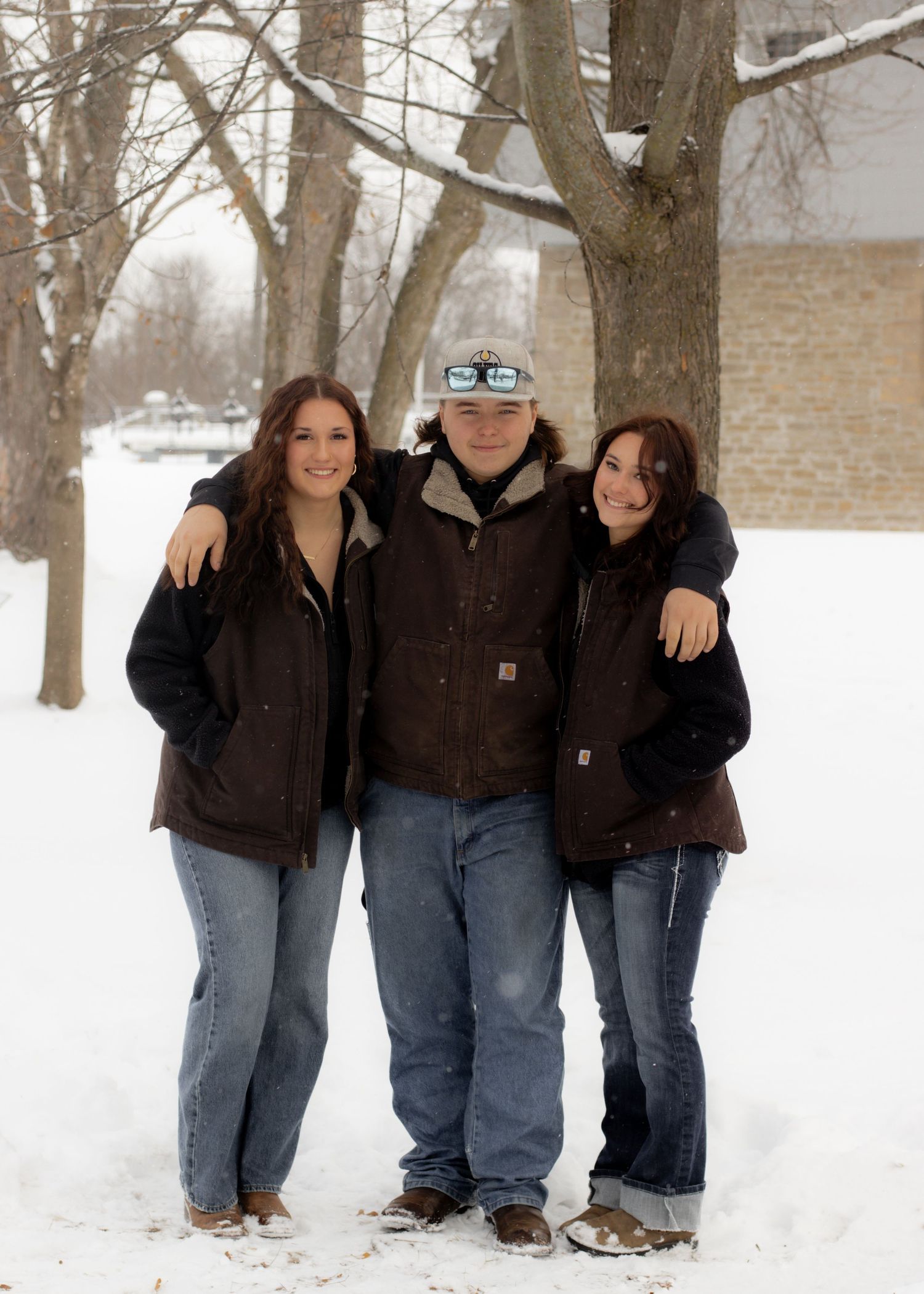 Three siblings posing together outdoors in matching brown jackets during winter snowfall.