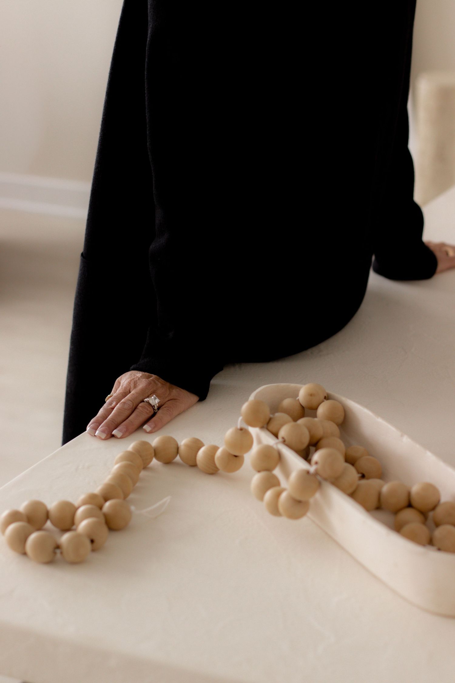 Woman in black outfit kneeling beside a white surface with decorative wooden bead garland.