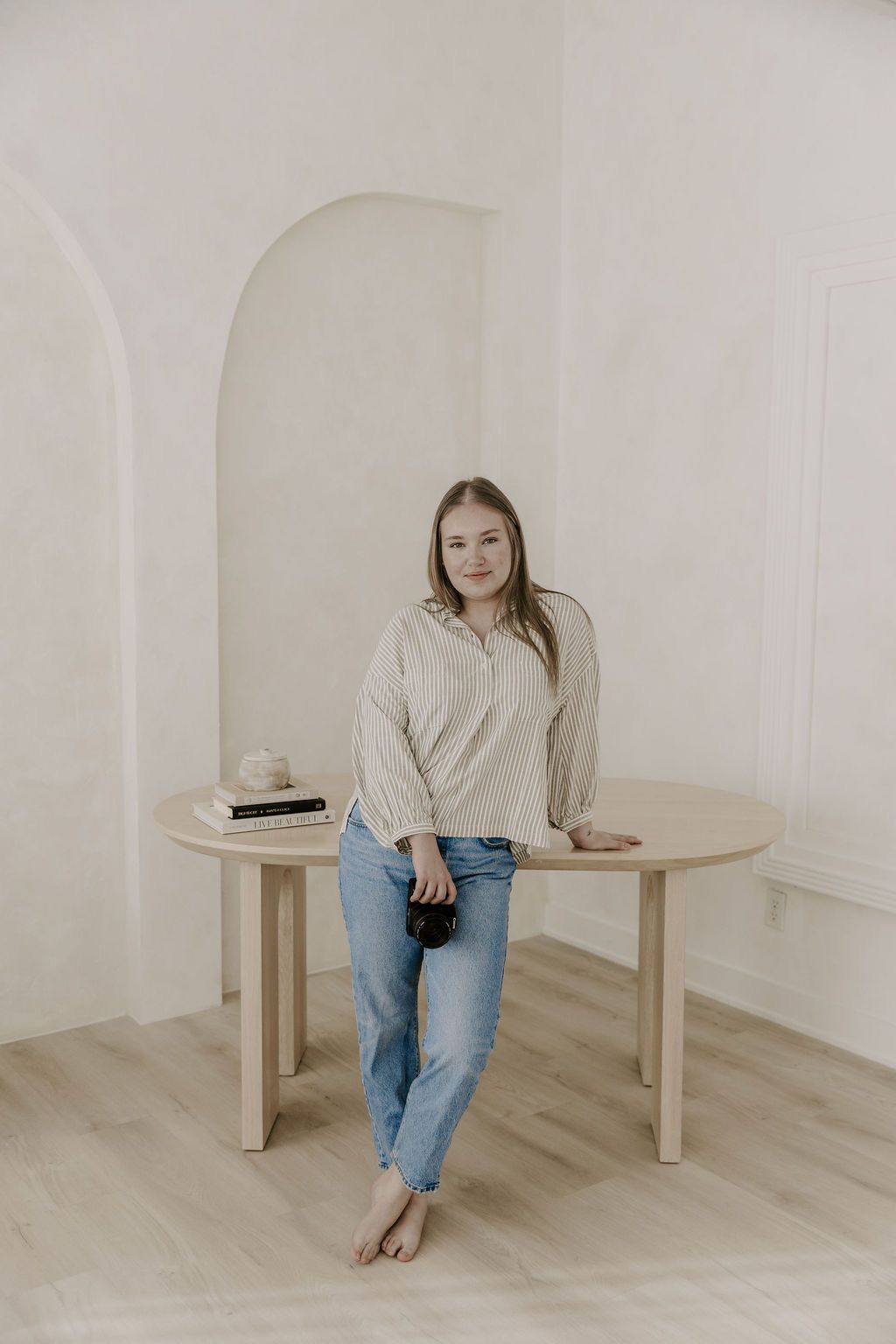 Smiling blonde woman in beige shirt and jeans leans against a round wooden table in a bright room.