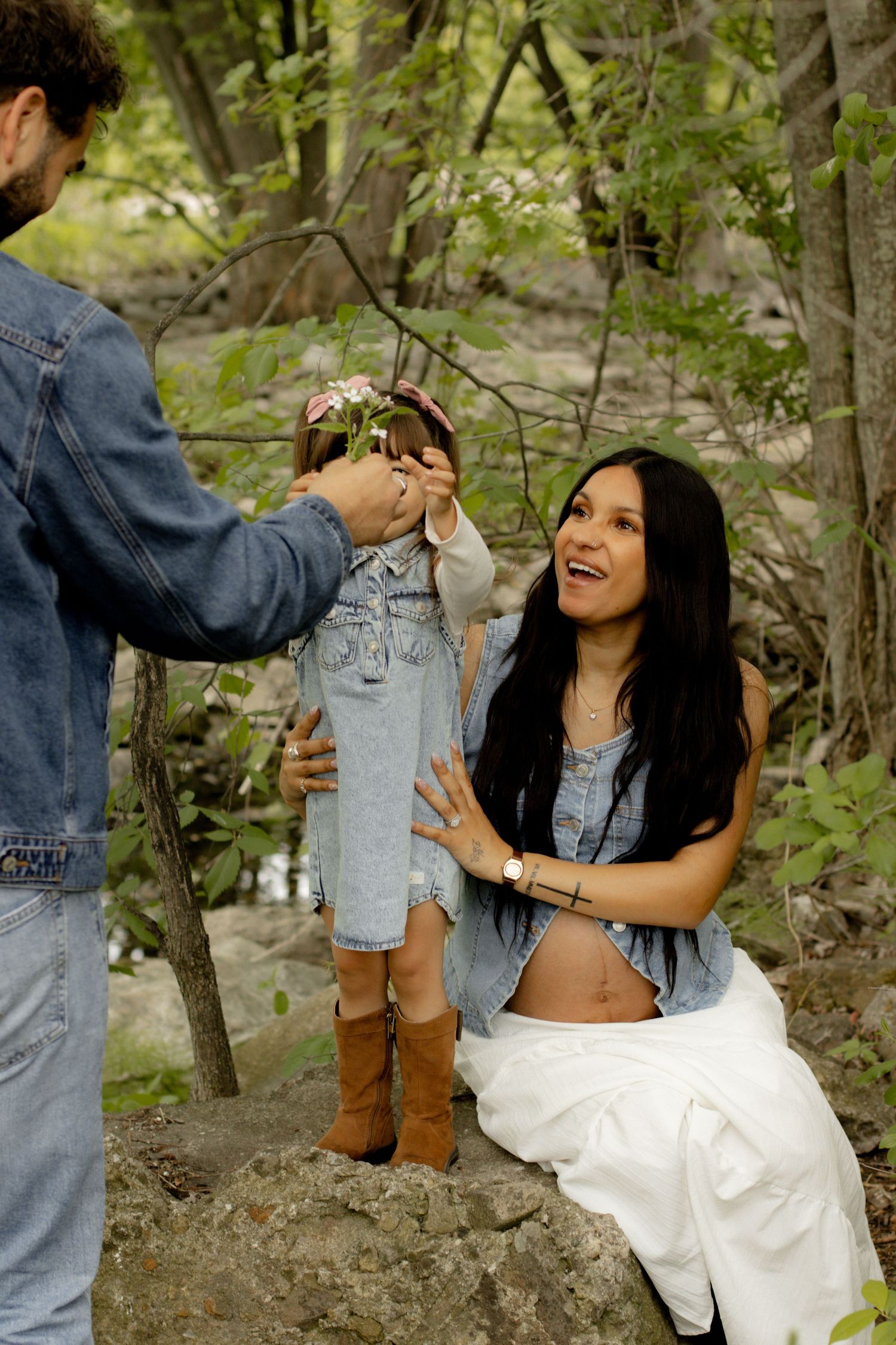 Pregnant woman smiling at toddler girl holding flowers, with man in denim jacket, outdoors in wooded setting.