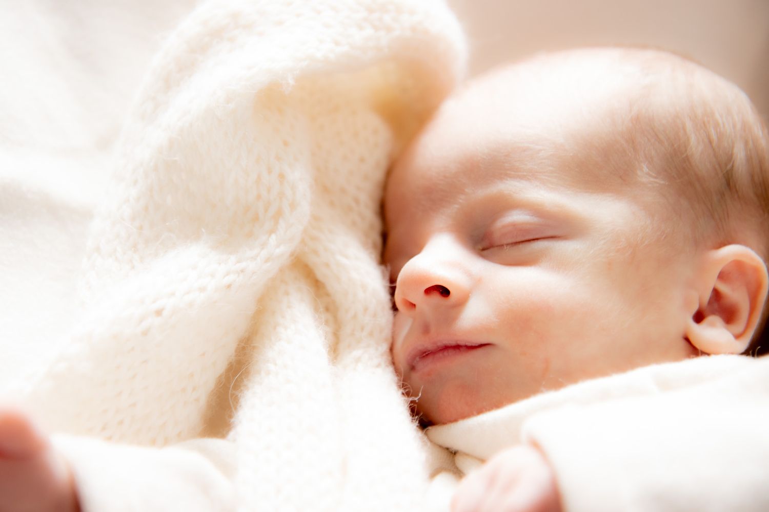 Peaceful newborn baby sleeping wrapped in a soft white knit blanket with eyes closed.