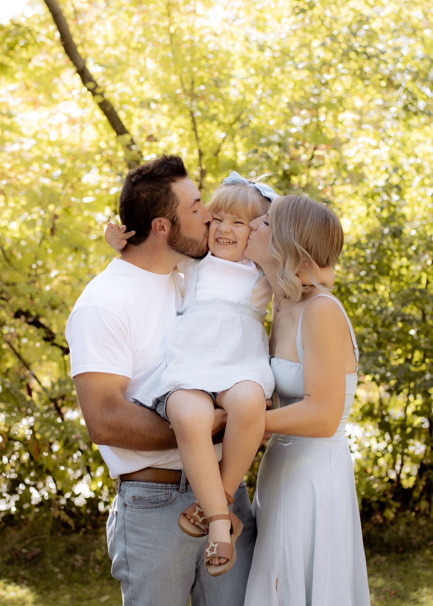 Parents kissing young toddler daughter on cheeks simultaneously while holding her outdoors.