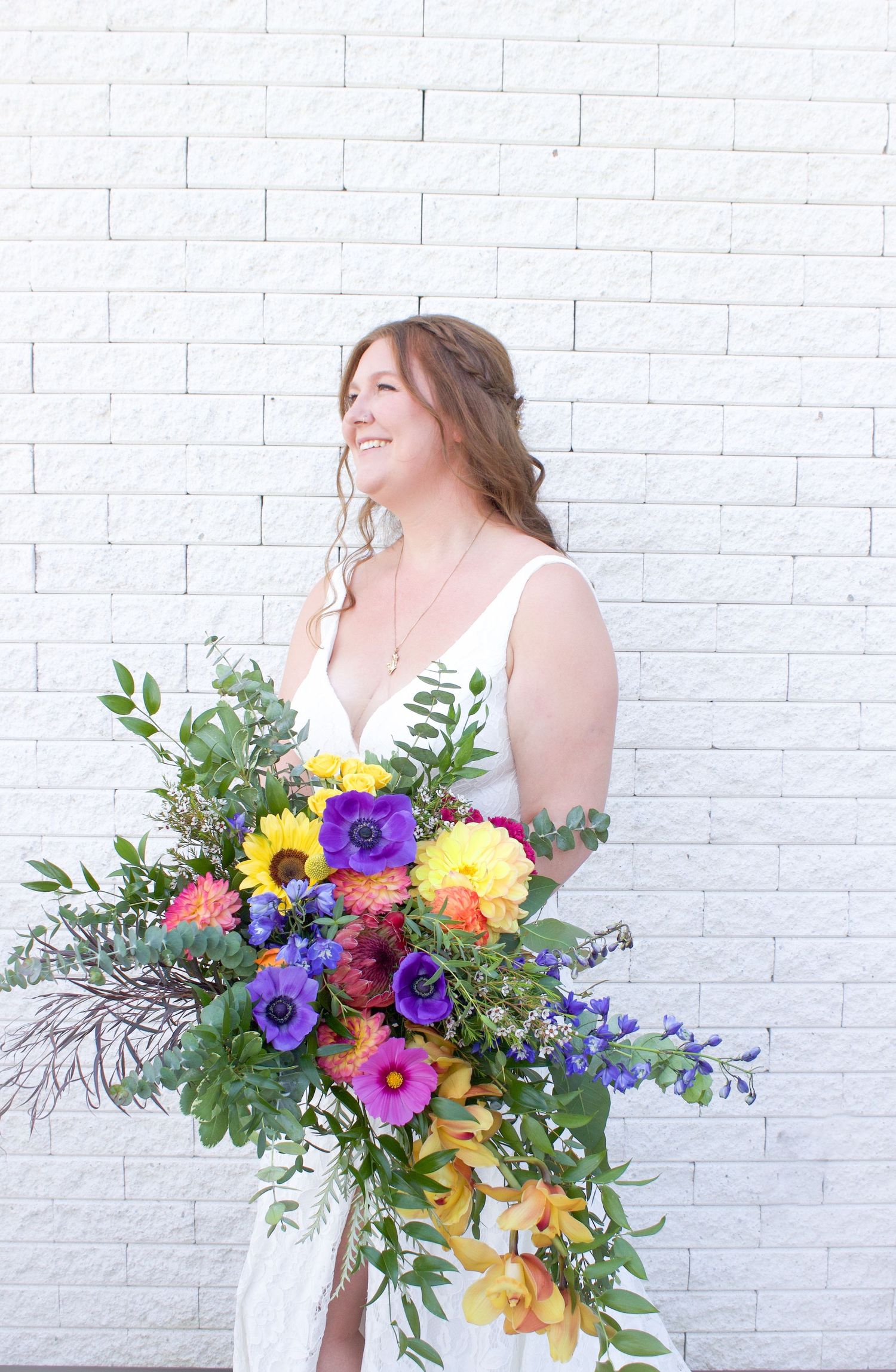 A joyful bride in a white gown holding a vibrant cascading bouquet with sunflowers and purple anemones.