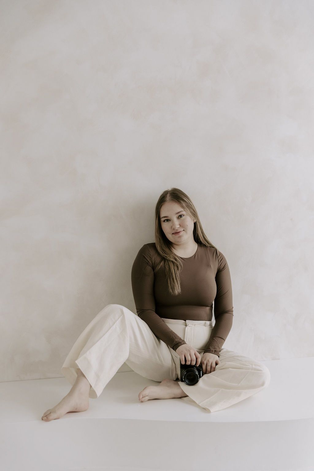Young woman in brown crop top and white pants sits cross-legged holding a camera against a neutral background.