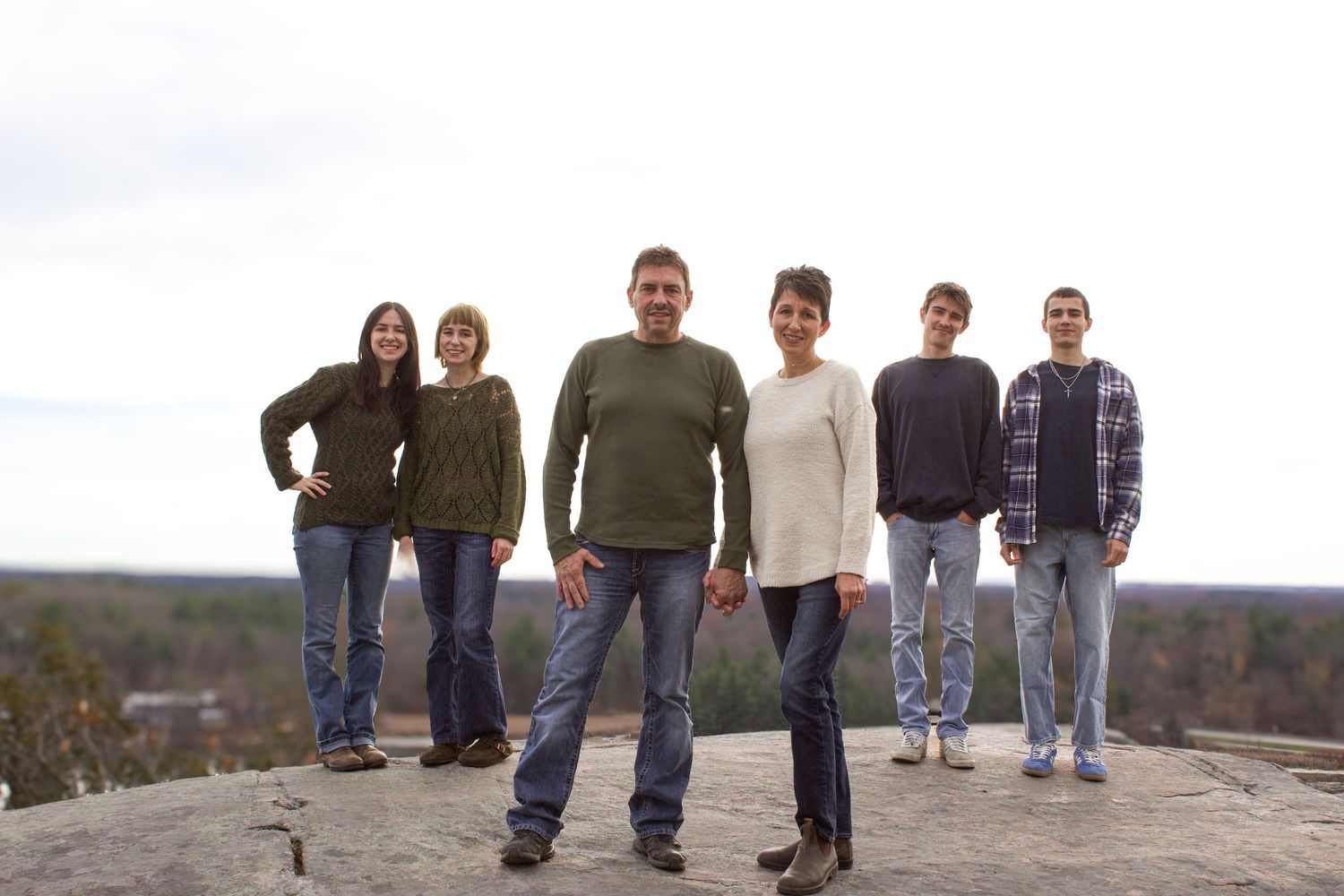 Family of six poses on rocky hilltop with sweeping autumn landscape, wearing coordinated earth-tone outfits.