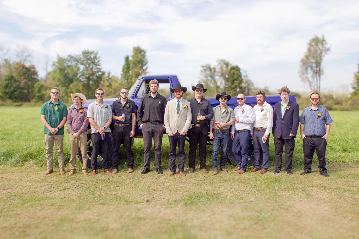 Group of groomsmen and wedding party posing outdoors in field near blue truck.